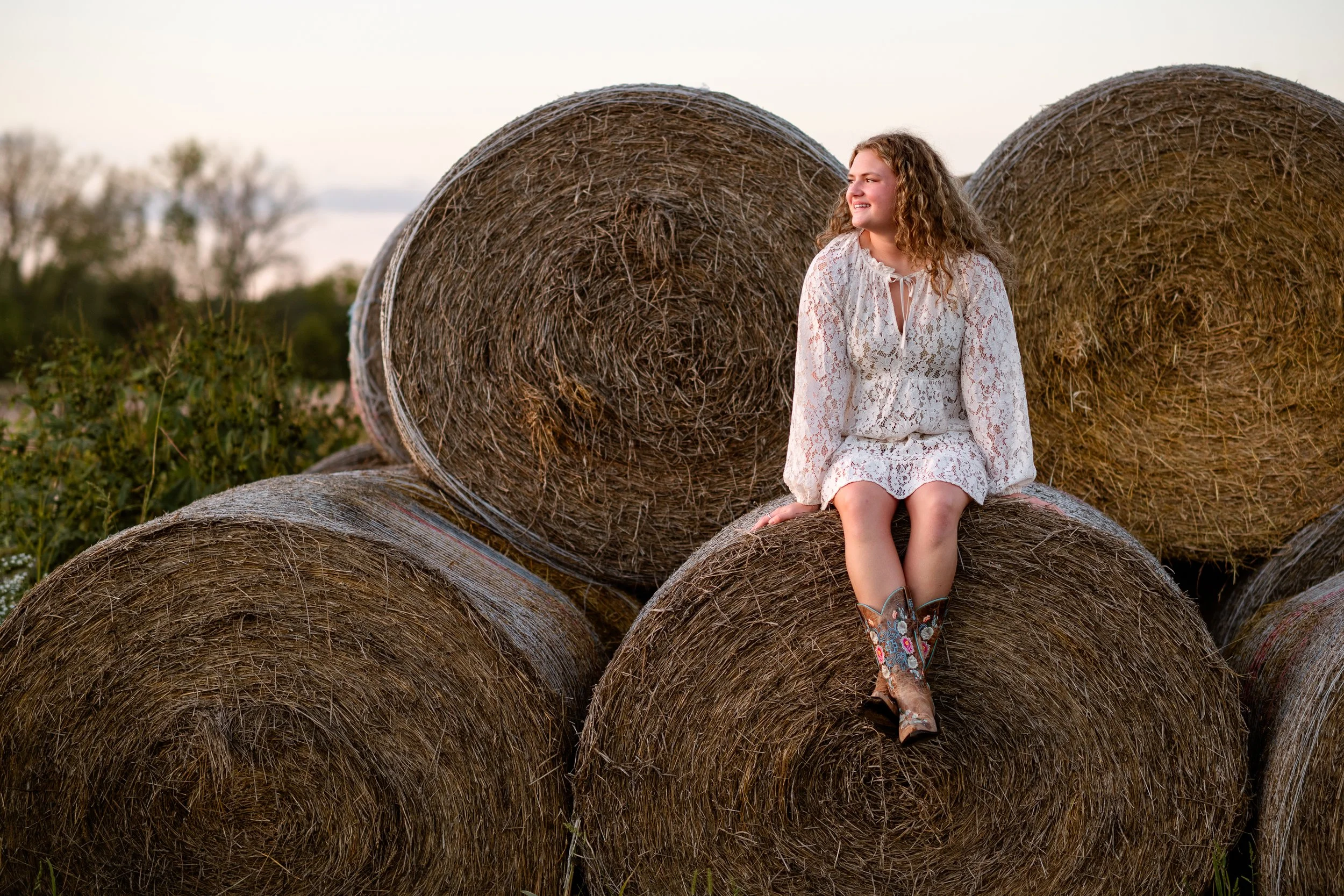 A young woman with curly hair wearing a white lace dress and cowboy boots sitting on a hay bale in a field with several hay bales around her during sunset.