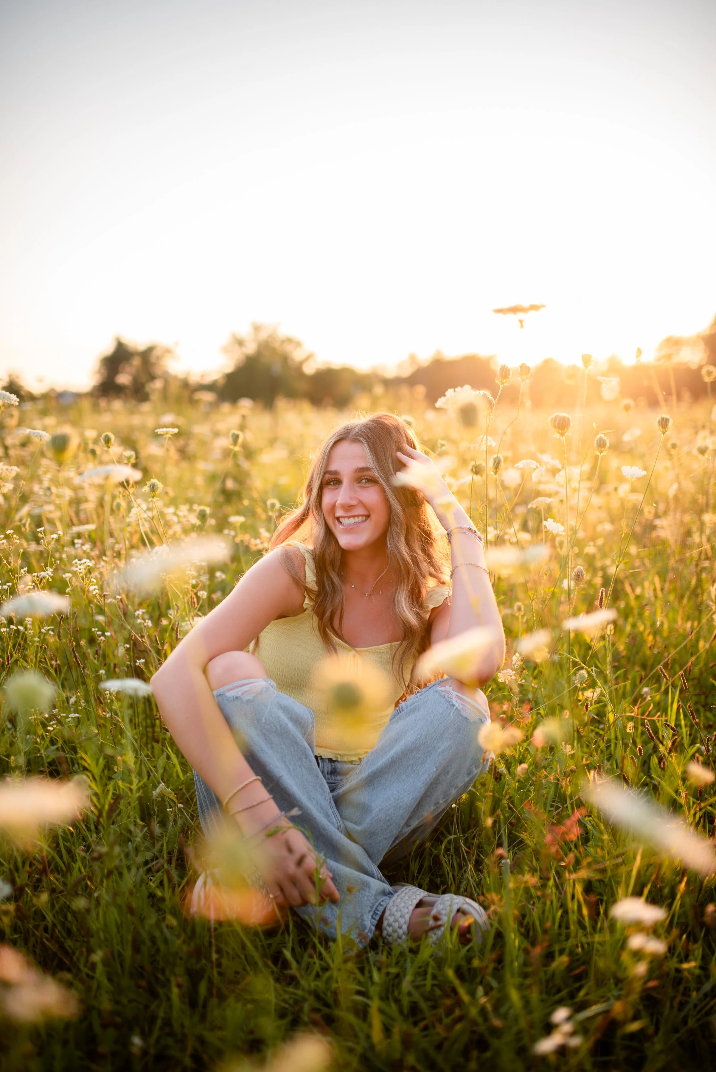 A young woman sitting in a field of yellow and white flowers during sunset, smiling and looking relaxed.