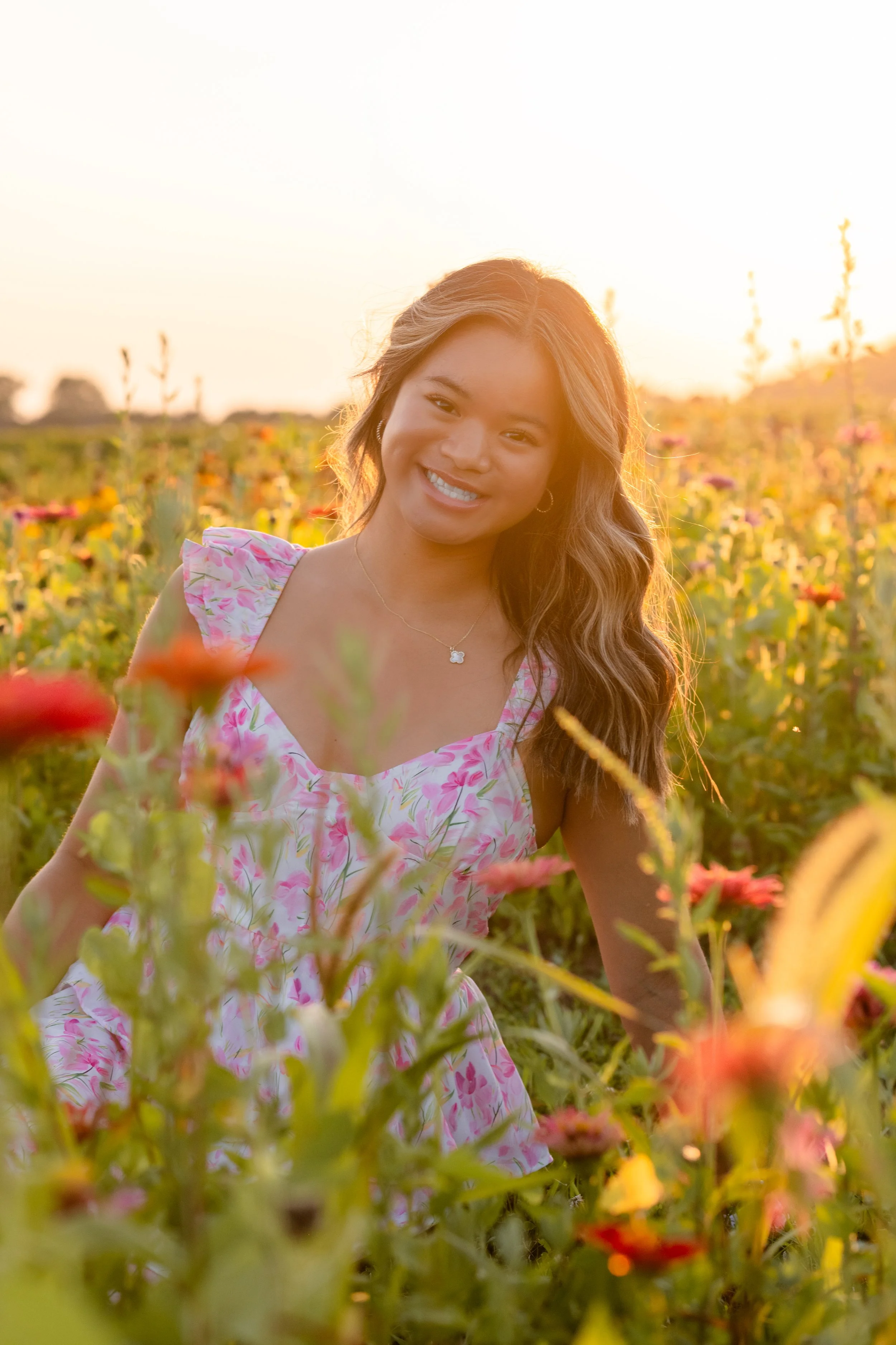 A woman with long wavy hair smiling in a field of colorful flowers during sunset.