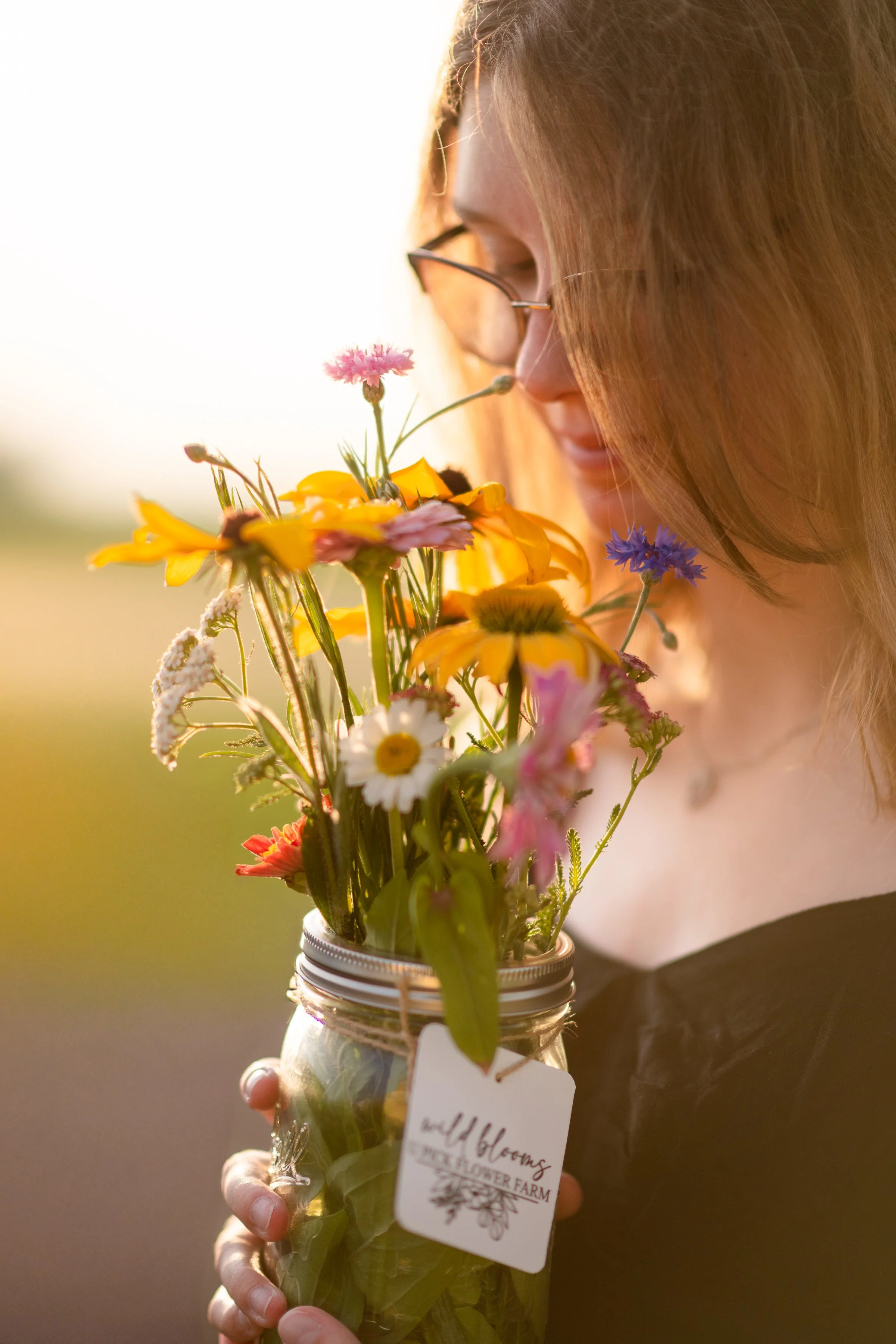 Person holding a glass jar filled with a colorful arrangement of wildflowers, including pink, yellow, white, and purple flowers, outdoors in warm lighting.