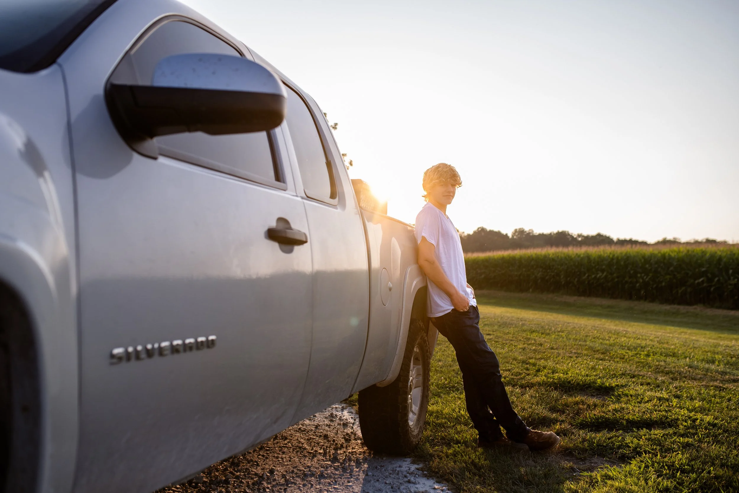 A young man with curly hair wearing a white t-shirt and jeans leaning against a silver Chevrolet Silverado pickup truck in a countryside setting during sunset.