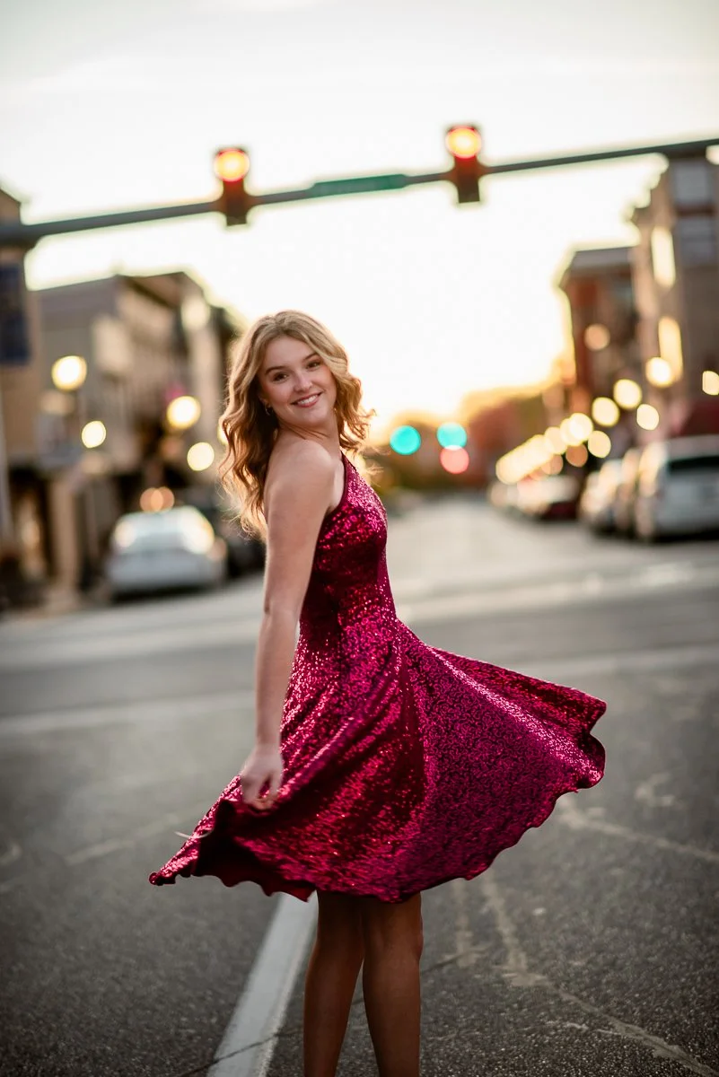 A young woman in a sparkling pink dress spins on a city street at sunset, with traffic lights and blurred parked cars in the background.