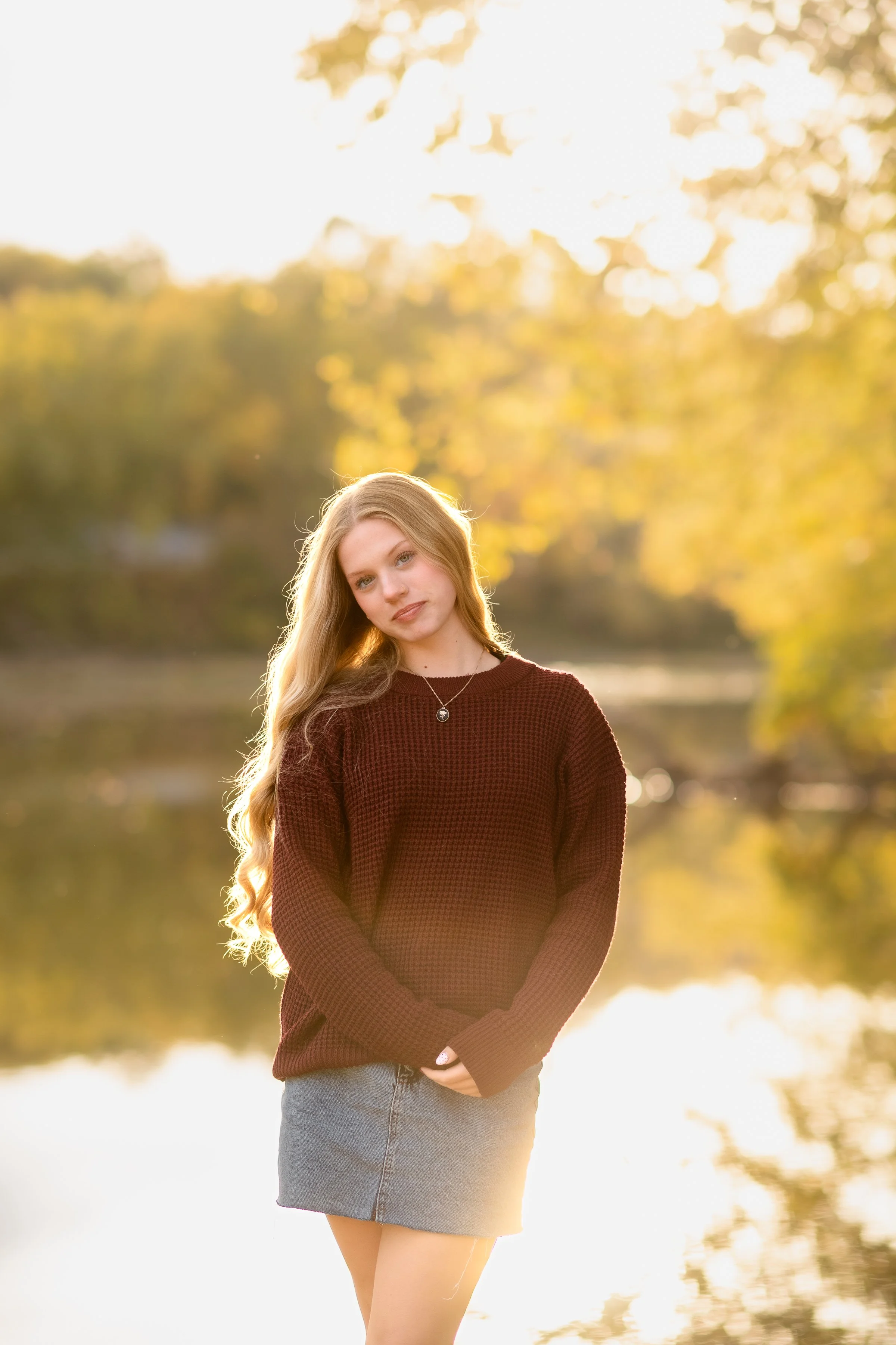 A young woman with long blonde hair standing outdoors during golden hour, wearing a maroon sweater and a denim skirt, with trees and water in the background.