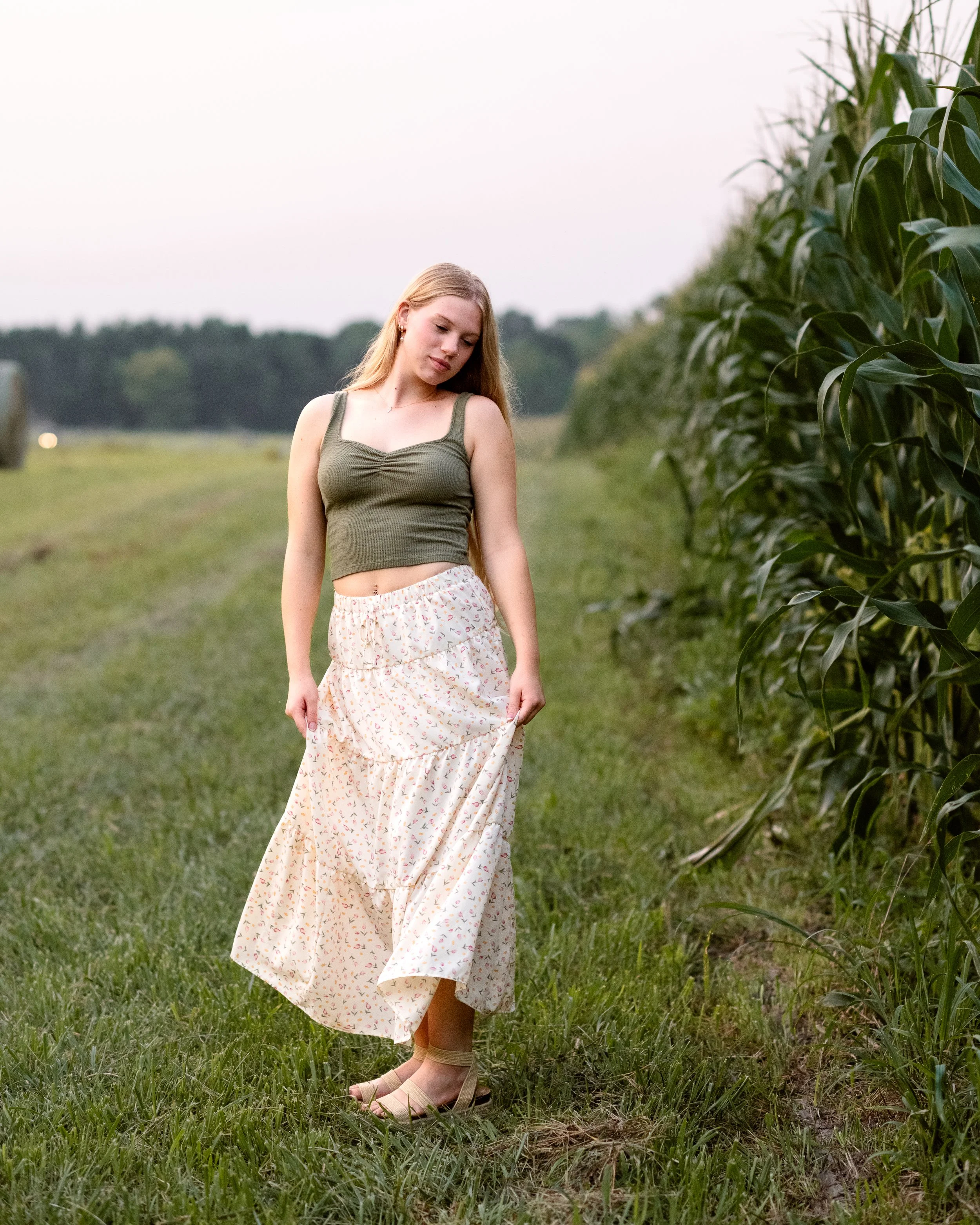 A young woman with long blonde hair standing outdoors near a row of tall green corn plants in a field, wearing a green tank top, a long floral skirt, and beige sandals, looking downward with a peaceful expression.
