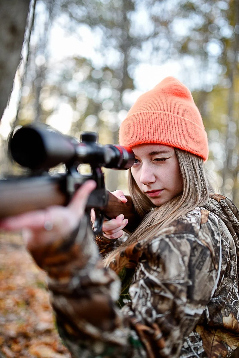 Young girl in camouflage jacket looking through a rifle scope outdoors in a forest with autumn foliage, wearing an orange beanie.