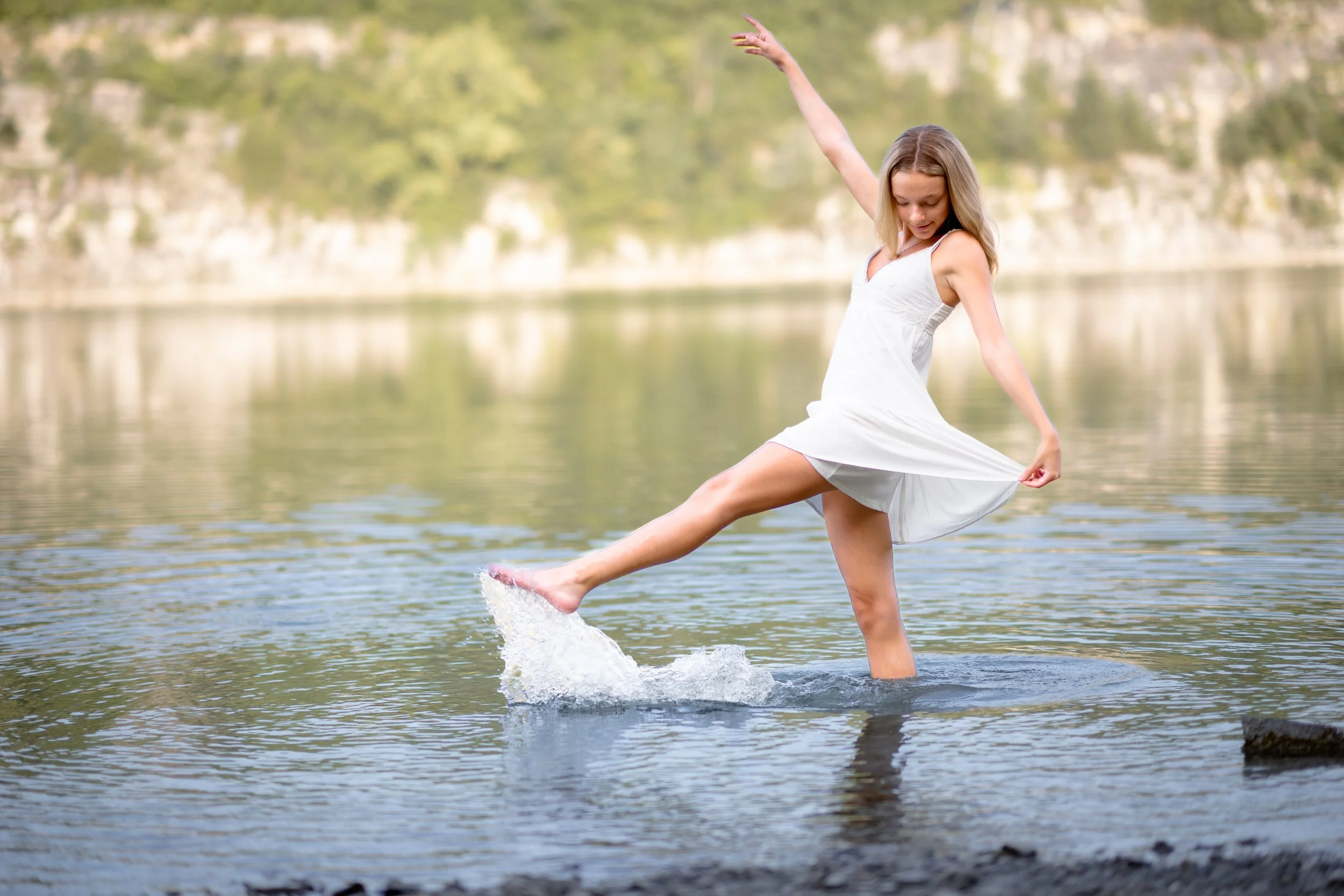 A young woman in a white dress balancing on a small rock in a body of water, lifting one arm and holding the hem of her dress with the other, with a blurred background of trees and cliffs.