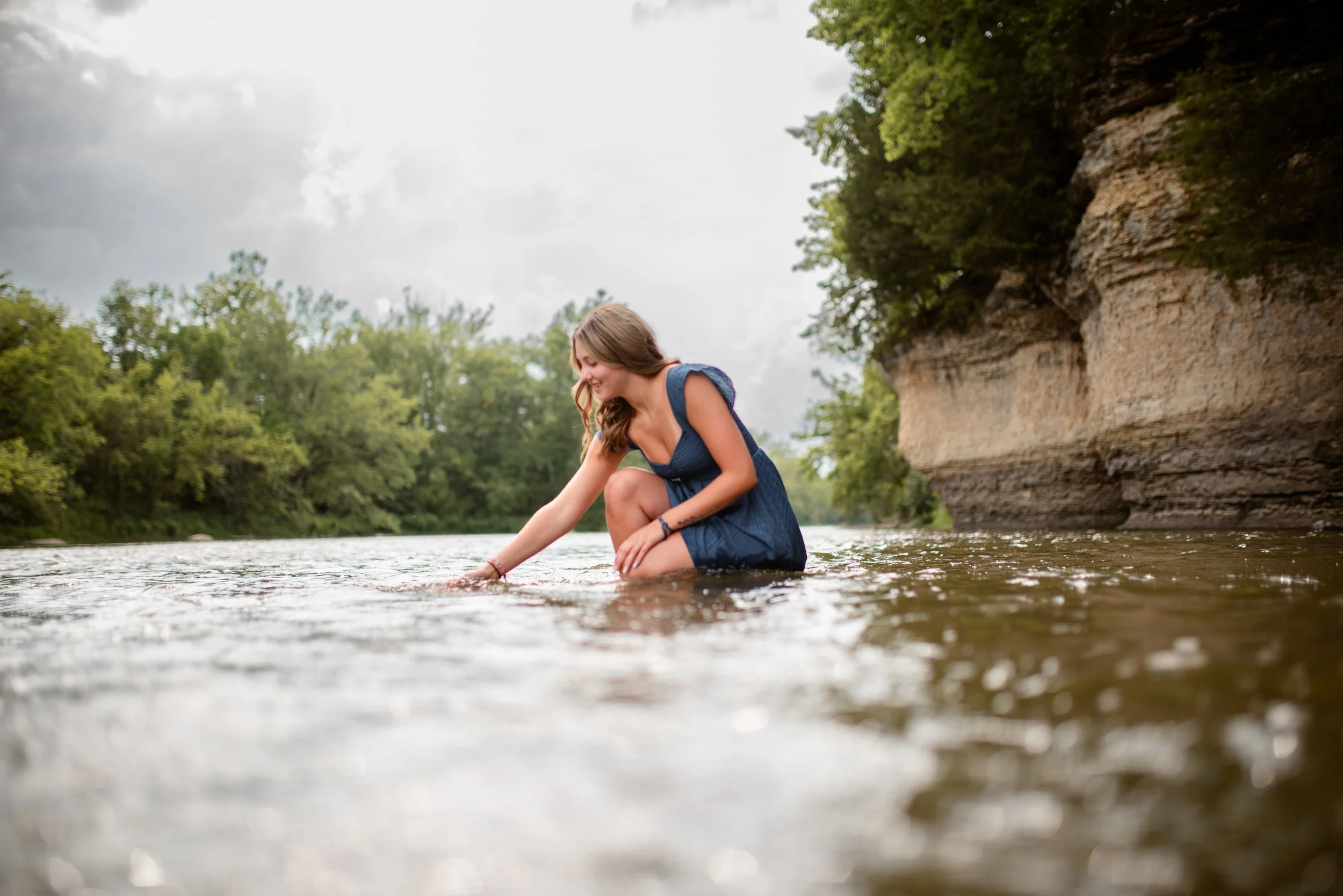 A young woman in a blue dress crouches in a river, smiling and touching the water near a cliff and green trees.