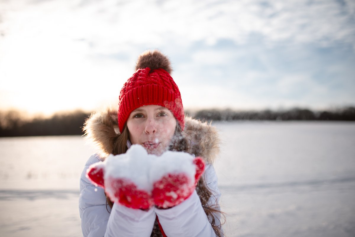 A woman outdoors in winter wearing a red knit hat with a pom-pom, a white jacket with a fur-lined hood, and red gloves, blowing snow from her hands.