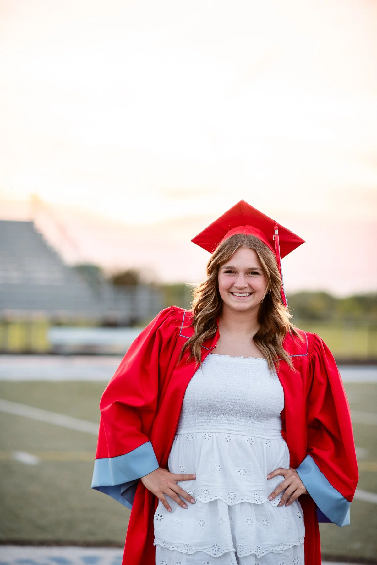 A young woman in a white dress and red graduation gown and cap standing on a football field during sunset.