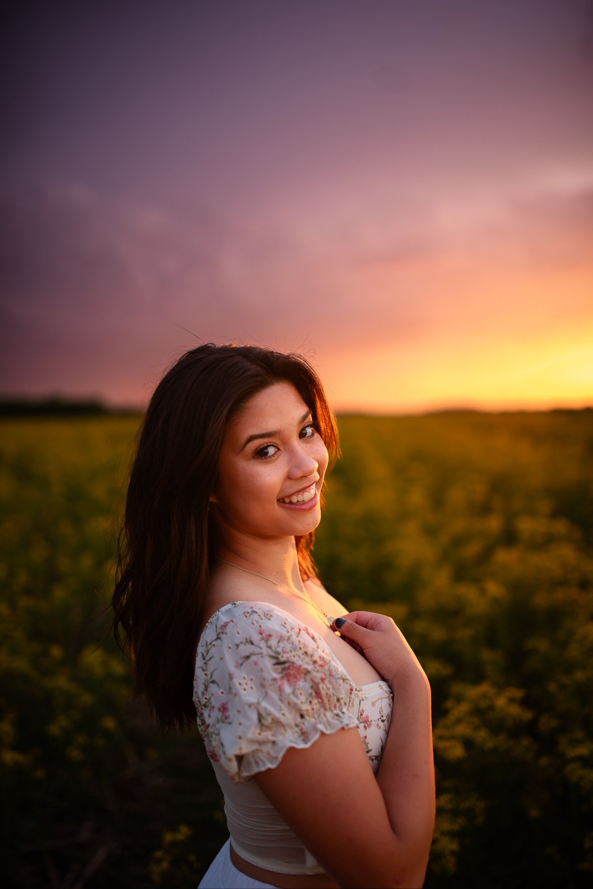 A young woman with dark hair smiling at sunset in a field, wearing a white floral dress.