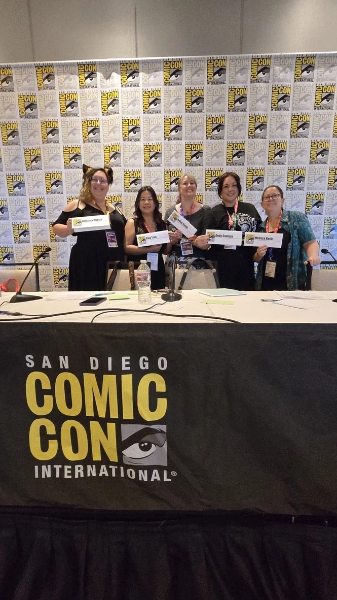 Five women standing and sitting behind a table at San Diego Comic-Con, holding name placards, with a backdrop of a Comic-Con logo wall.