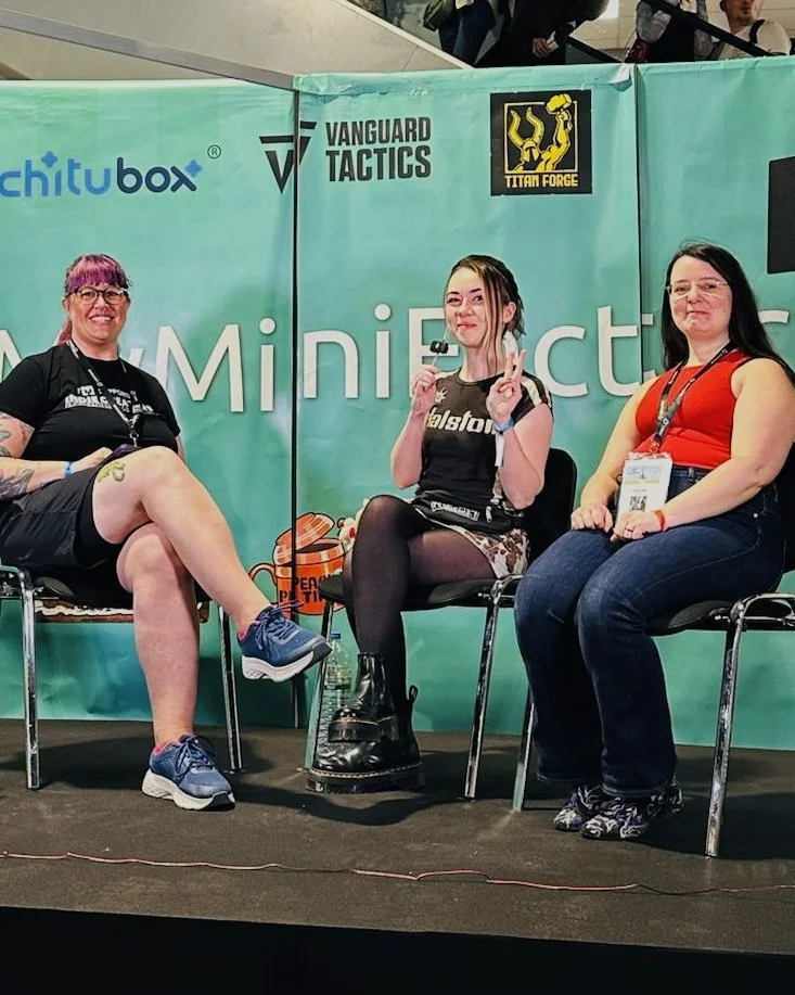 Three women sitting on chairs at a convention or expo, with a teal background featuring logos for 'Chitubox,' 'Vanguard Tactics,' and 'Titan Force.'