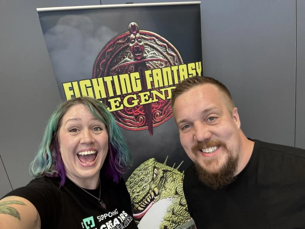 A woman with colorful hair and a man with a beard smiling in front of a Fighting Fantasy Legends banner.