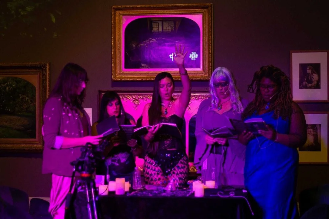 Five women standing around a table with lit candles, reading from books in a dimly lit room with framed artwork on the wall, illuminated by purple lighting.