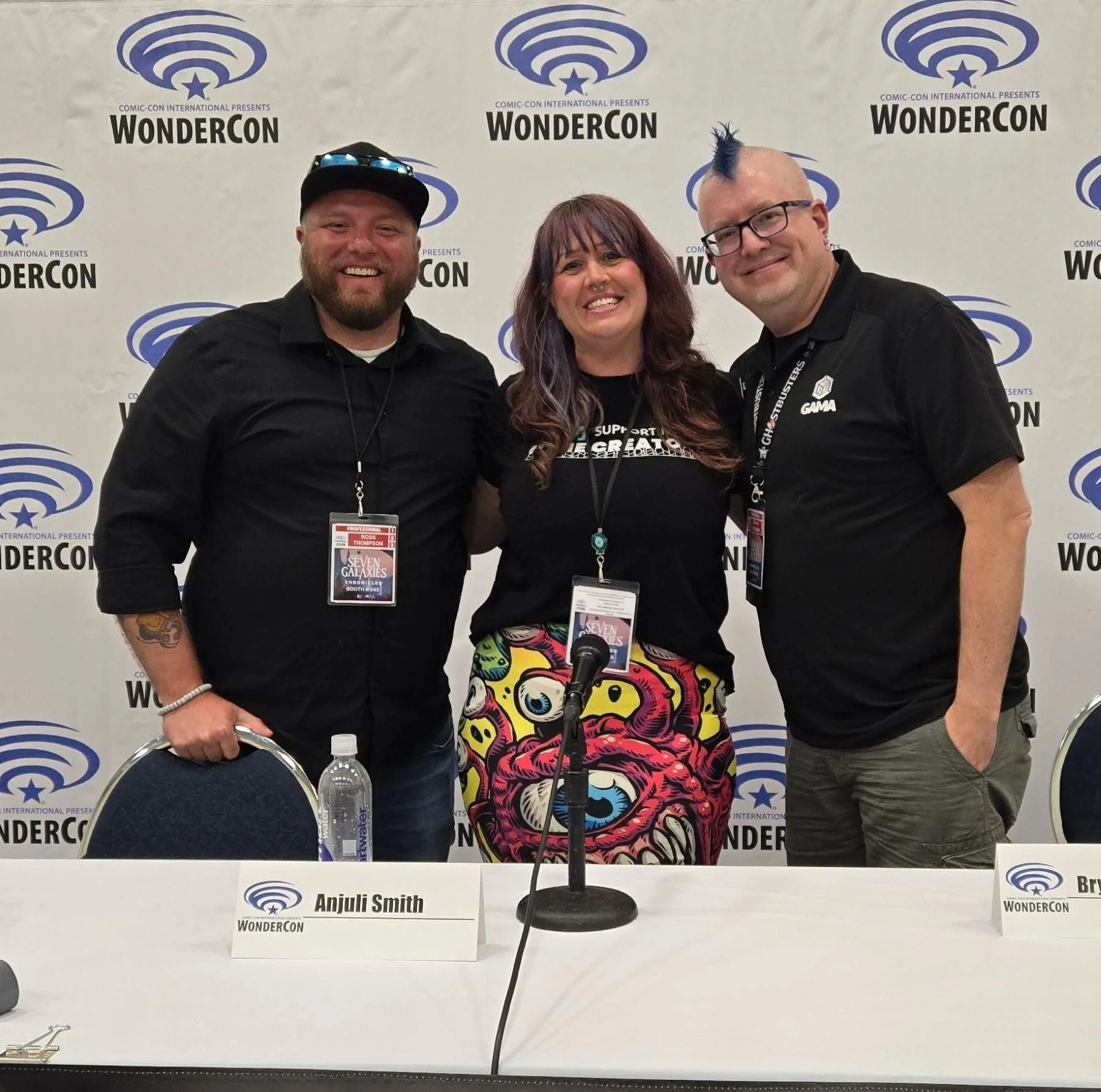 Three people smiling at a panel event with WonderCon logos in the background. The person on the left is a man with a beard, wearing a black shirt and a black cap with sunglasses on it. The person in the middle is a woman with long, wavy hair, wearing a black shirt and colorful pants with a skull pattern. The person on the right is a man with a mohawk, glasses, and a black shirt. There are name tags and a microphone on the table in front of them.