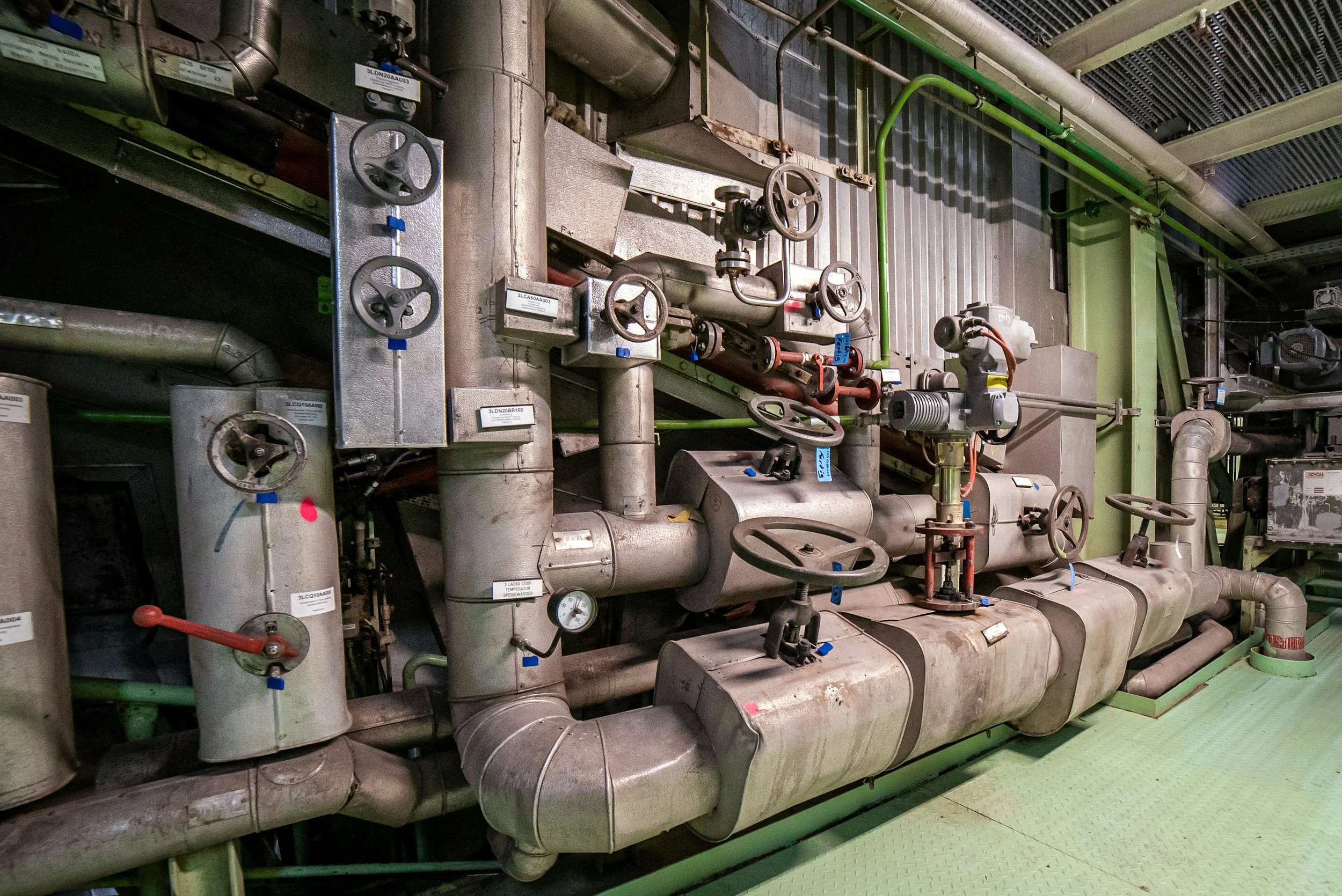Industrial piping system with various pipes, valves, and gauges in a factory or mechanical room.