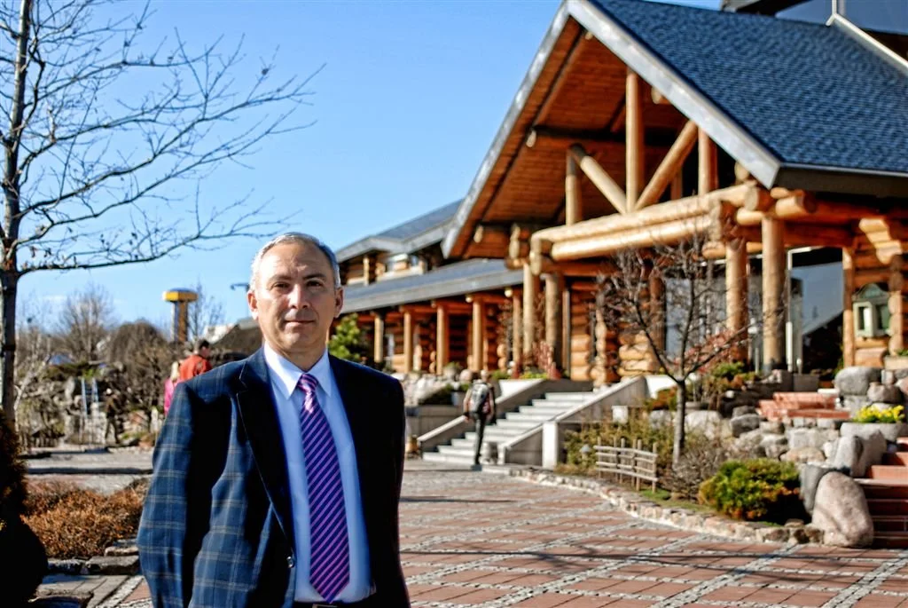 A man in a suit standing outdoors in front of a log cabin-style building on a sunny day.