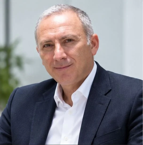 A middle-aged man with short gray hair in a navy suit and white shirt, sitting in an office or conference room.