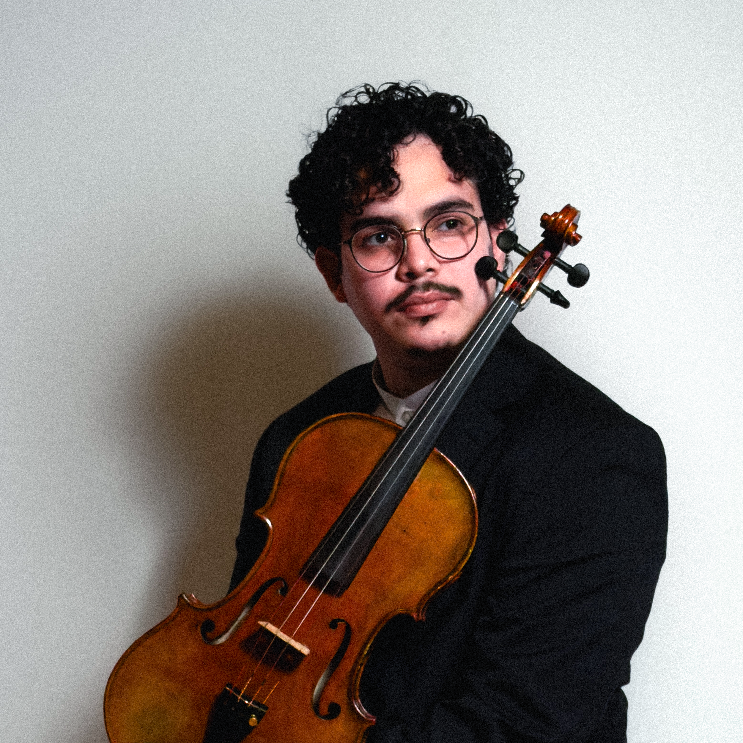 A person with curly dark hair and glasses holding a violin and bow against a plain light background.