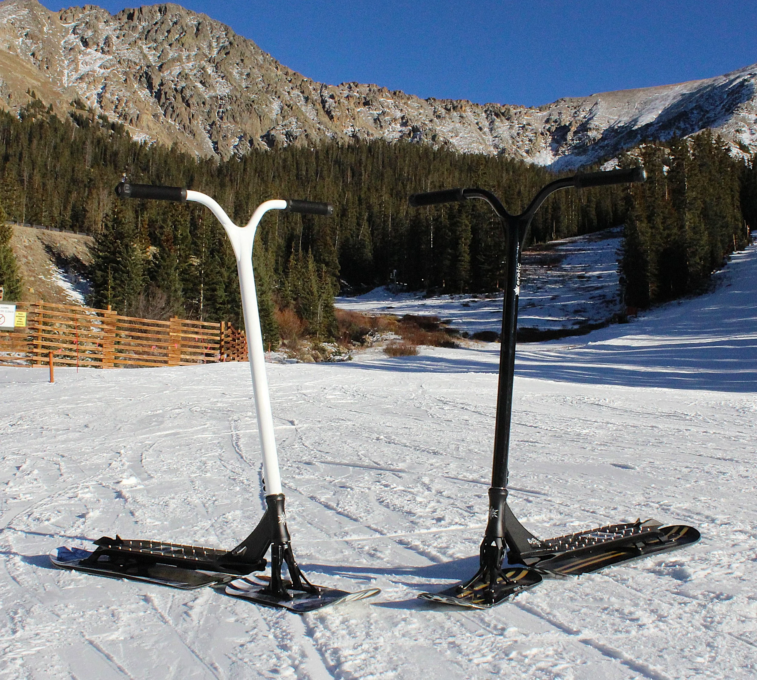 Two snow-covered eretic snowscooters in the foreground with a forested mountain slope and snow patches in the background. Clear blue sky overhead.