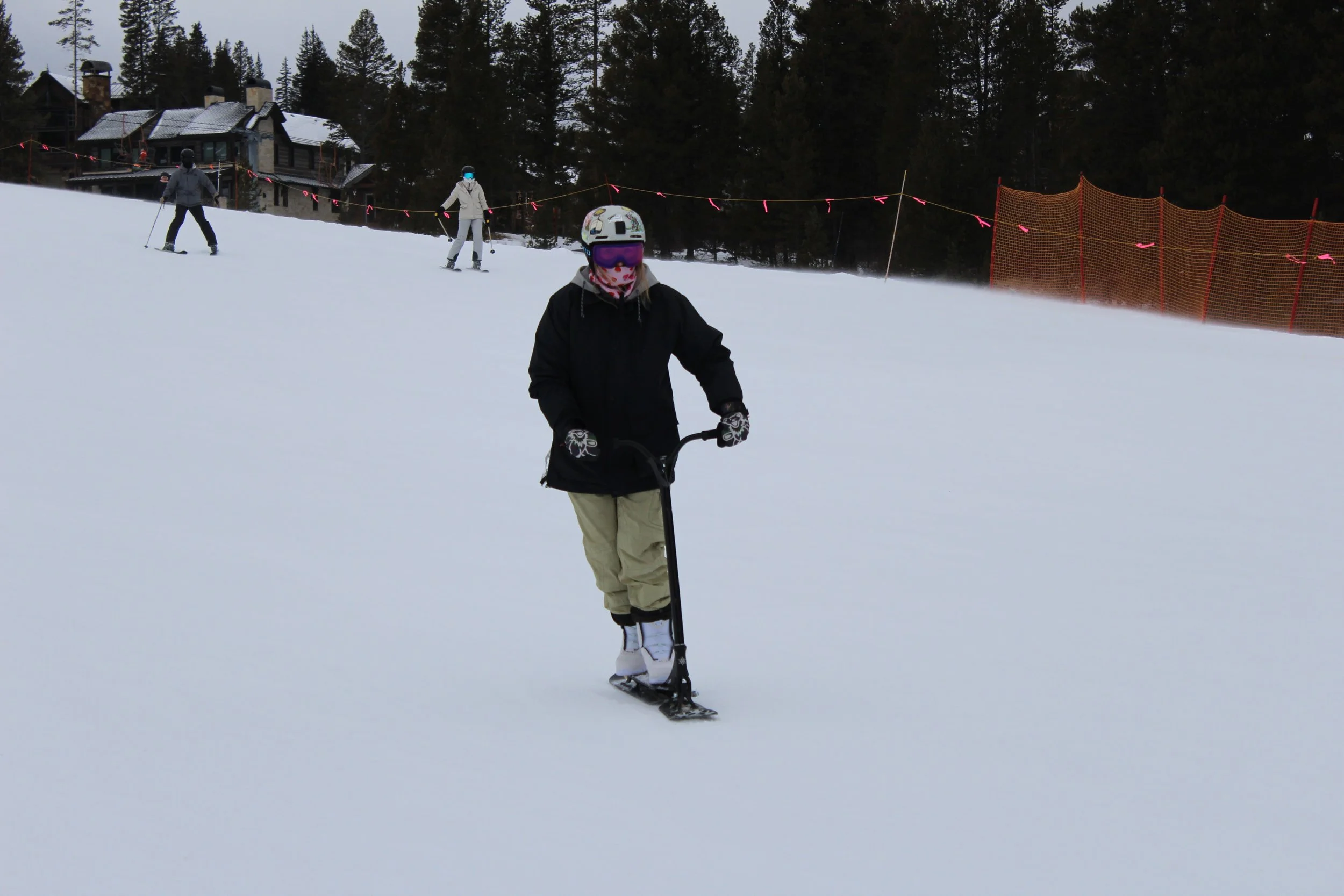 People skiing and snowboarding on a snowy slope near a forest and lodge buildings.