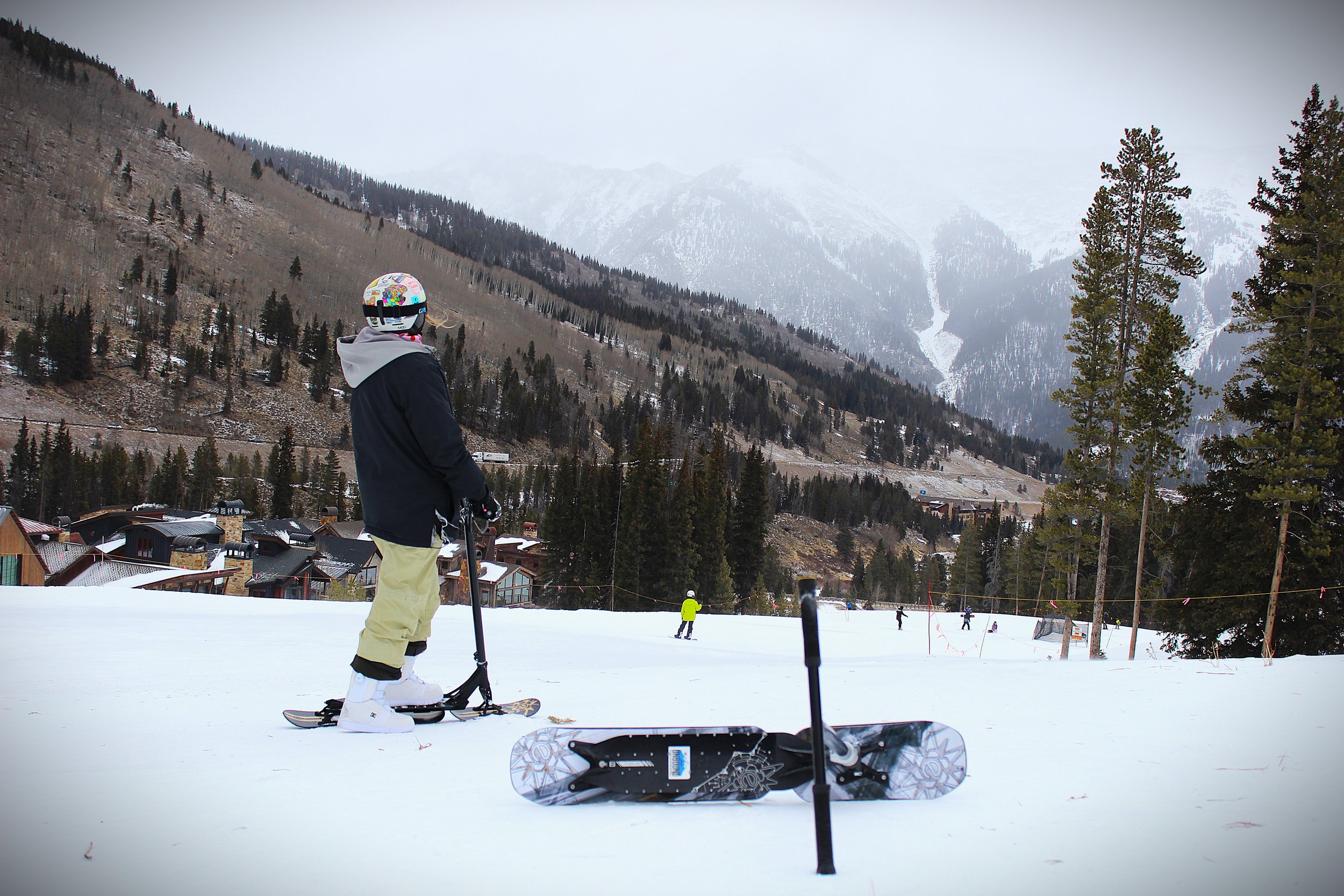 A person wearing winter clothing, including a helmet, standing on snow with snowscooters nearby, at a ski resort with trees, mountains, and buildings in the background.