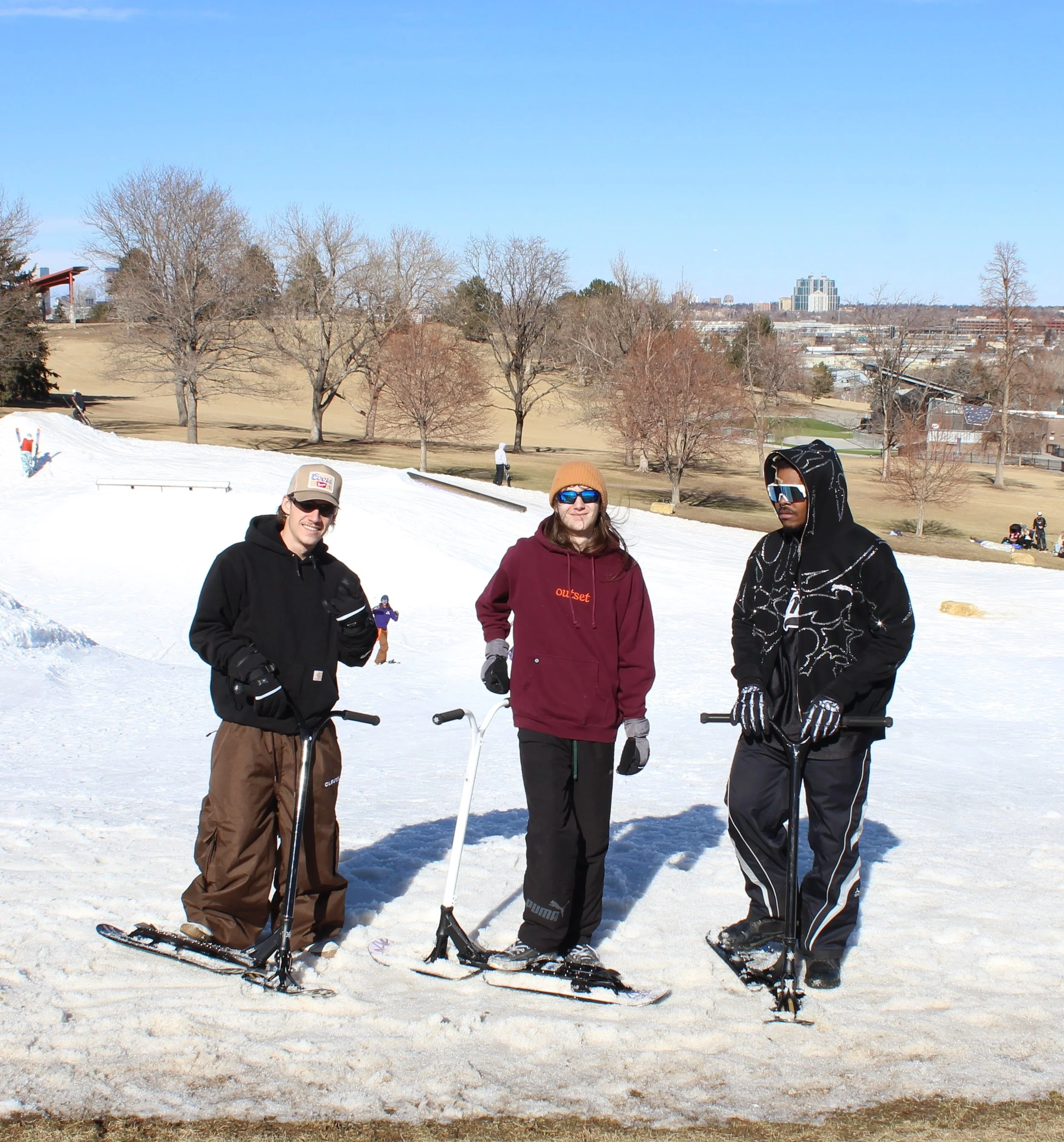 Three young men standing on snow-covered ground with snowscooter equipment, smiling in a terrain park with leafless trees and a cityscape background on a clear winter day.