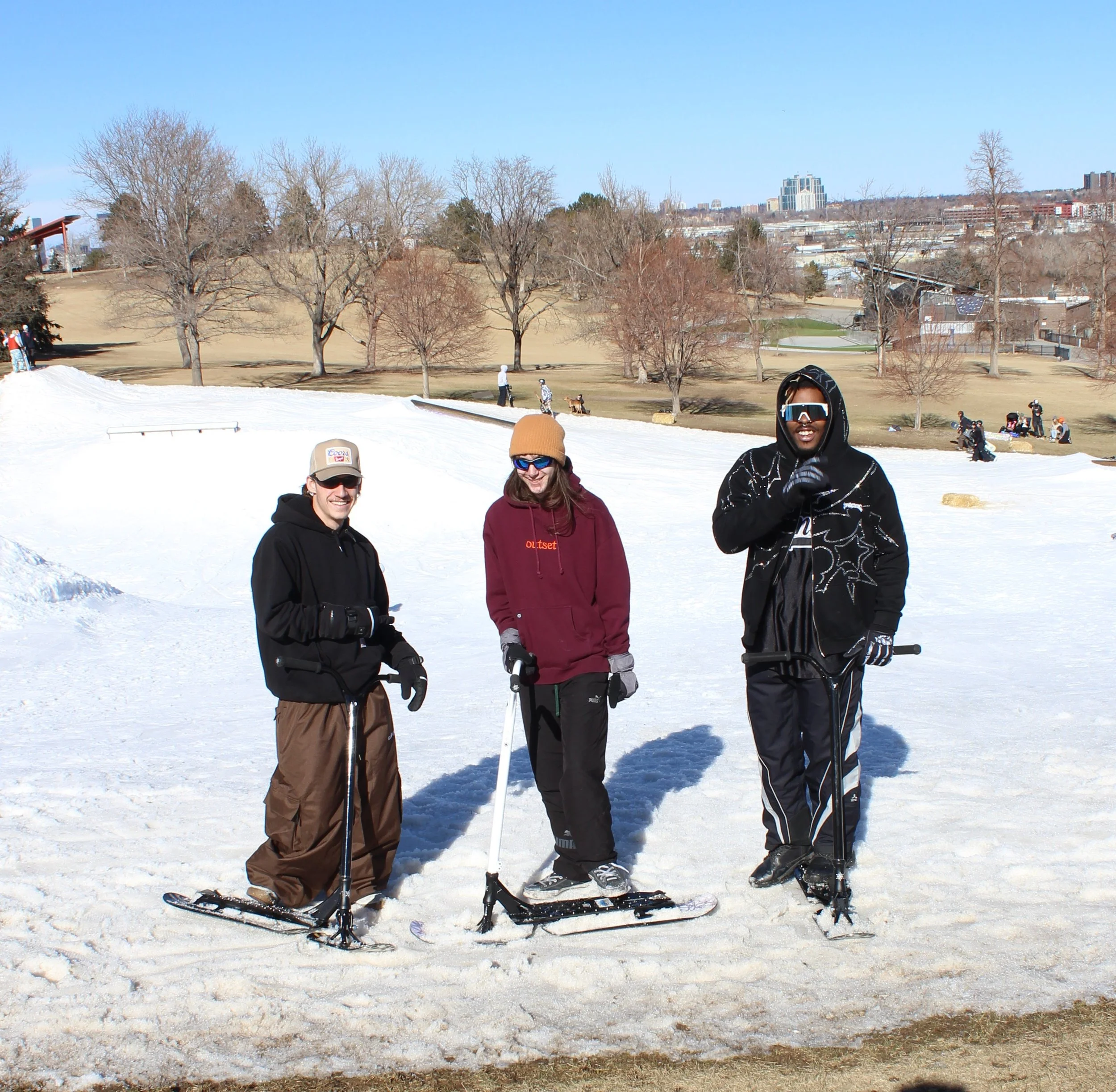 Three people standing with snow-covered ground in a park, holding snowboards and wearing winter clothing, with leafless trees and city buildings in the background.