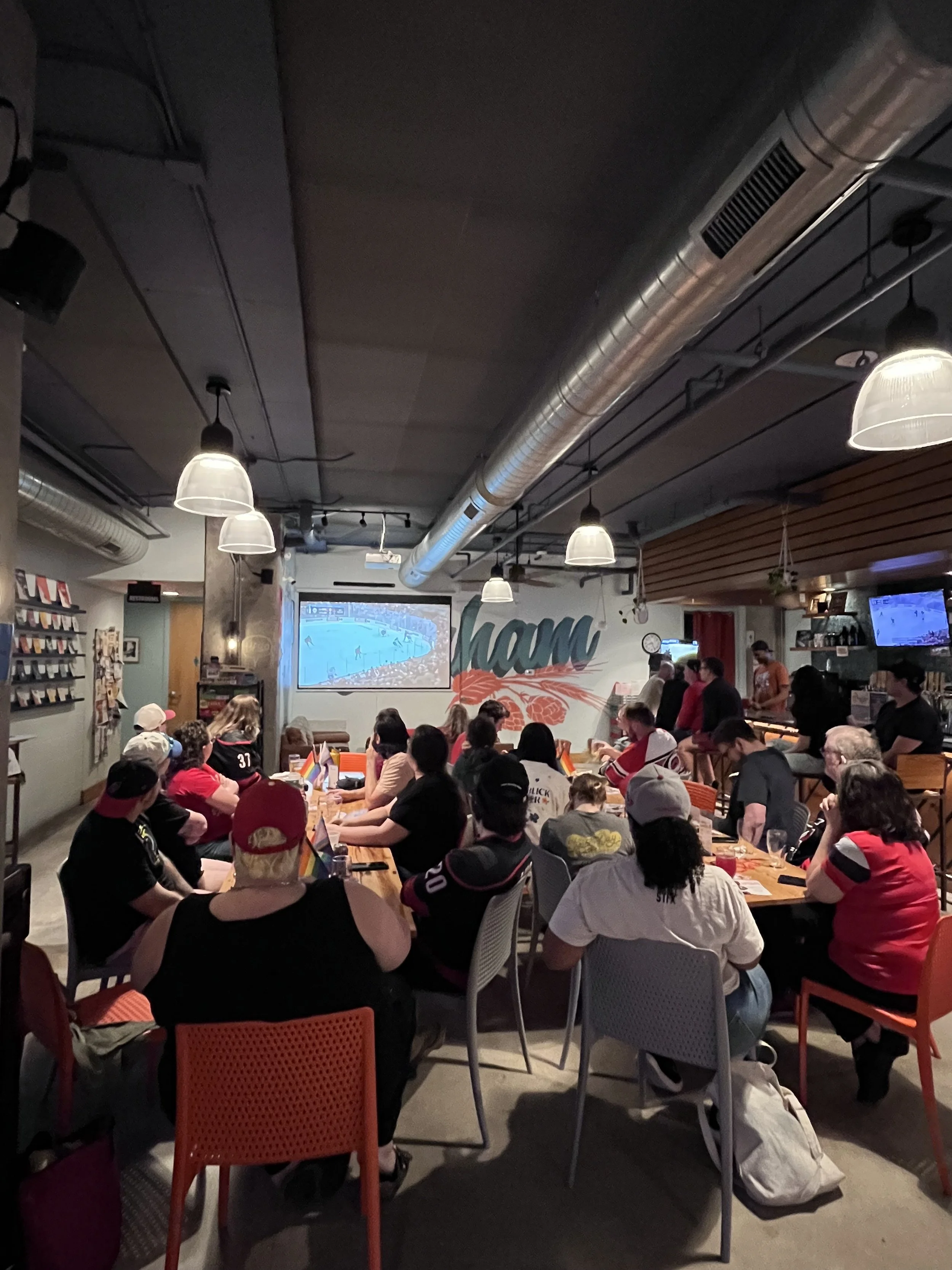 Indoor gathering at a brewery with people sitting at tables in a restaurant, with a large screen monitor showing a Carolina Hurricanes hockey game on the wall.
