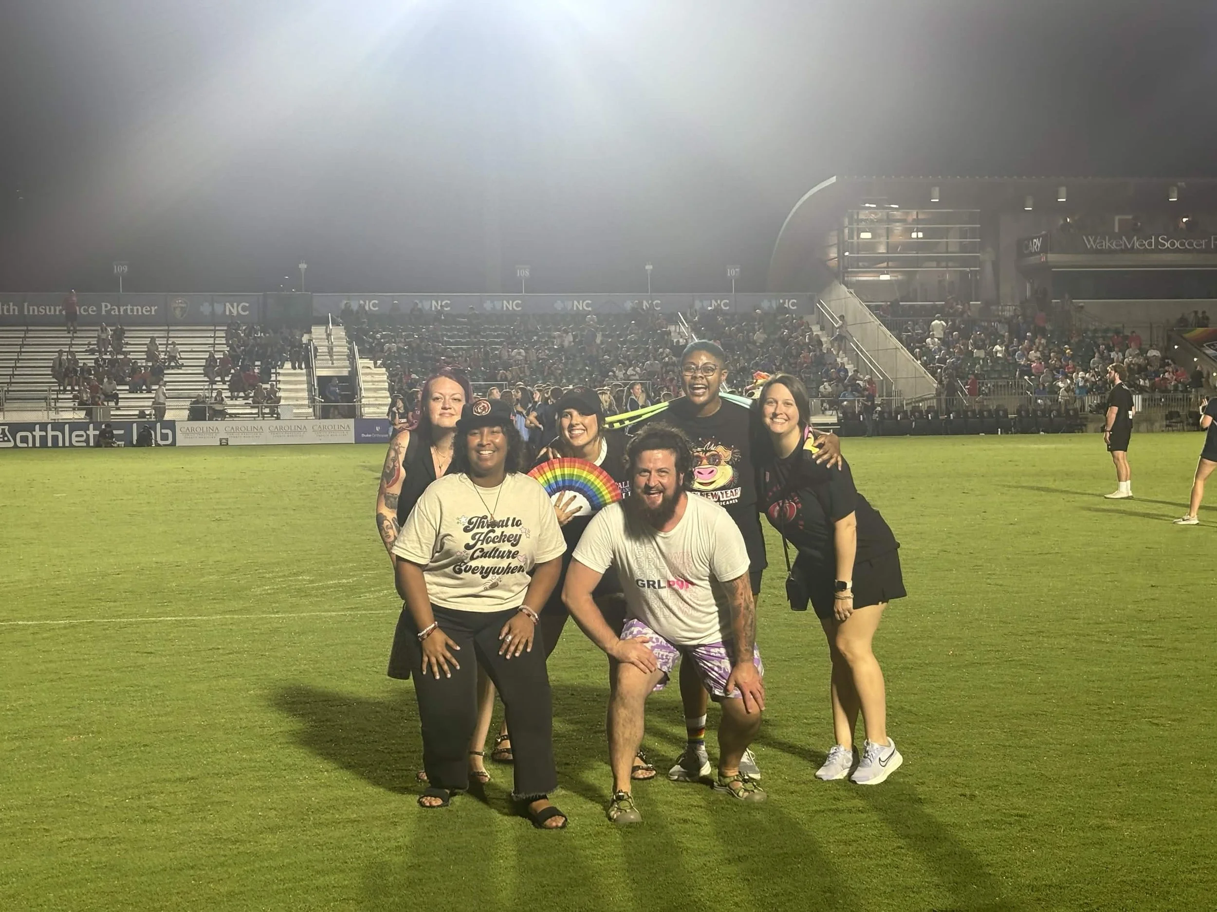A group of people standing together and smiling on a soccer field. Soccer stadium and fans can be seen in the background.