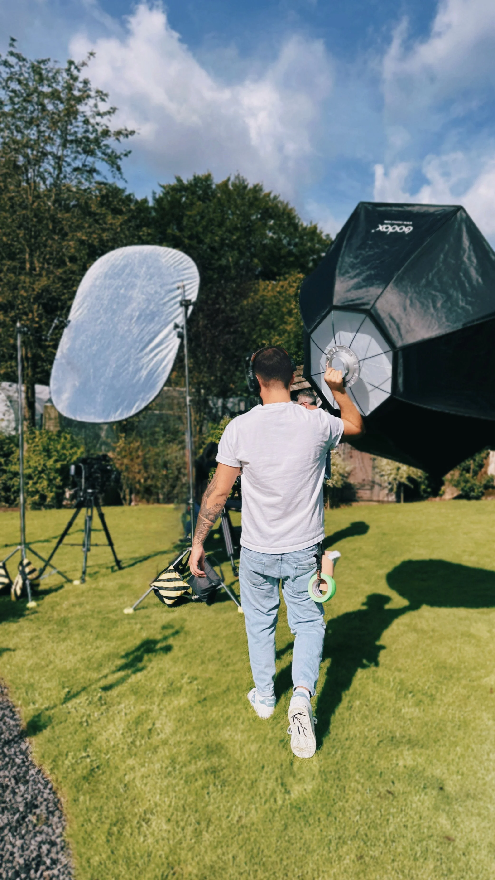 Ein junger Mann mit Tattoos auf dem linken Arm läuft auf einer Wiese während eines Fotoshootings im Freien. Im Hintergrund sind große Fotoblitze und Reflektoren zu sehen.
