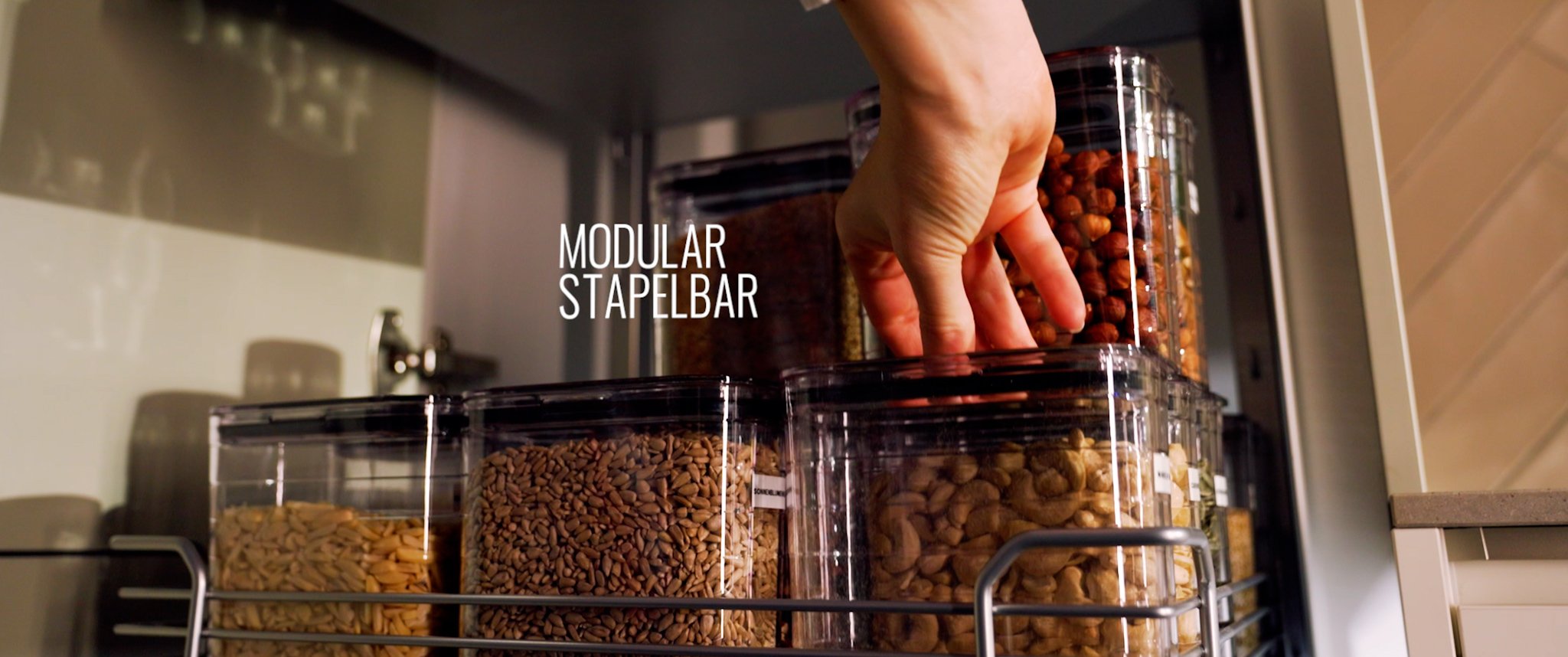 Hand greift in einen modularen Stapelbar-Behälter mit Nüssen, auf einem Regal mit mehreren transparenten Behältern voller Samen oder Nüsse