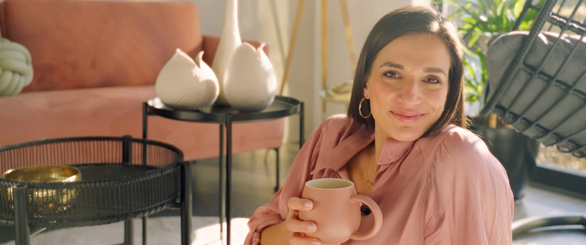 Eine lächelnde Frau mit braunen Haaren und goldenem Ohrring hält eine pinkfarbene Tasse in der Hand in einem schön dekorierten Raum.