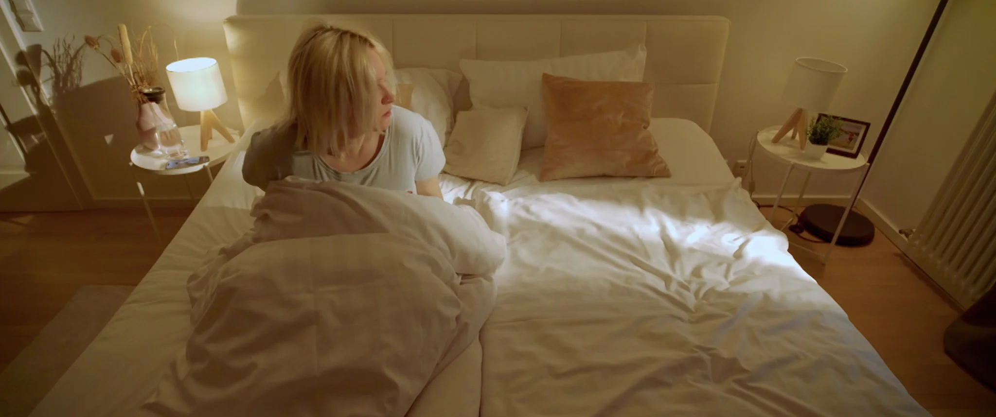 Cinematic SHort FIlm A woman sits covered up in bed, looking to the left in a bedroom.
