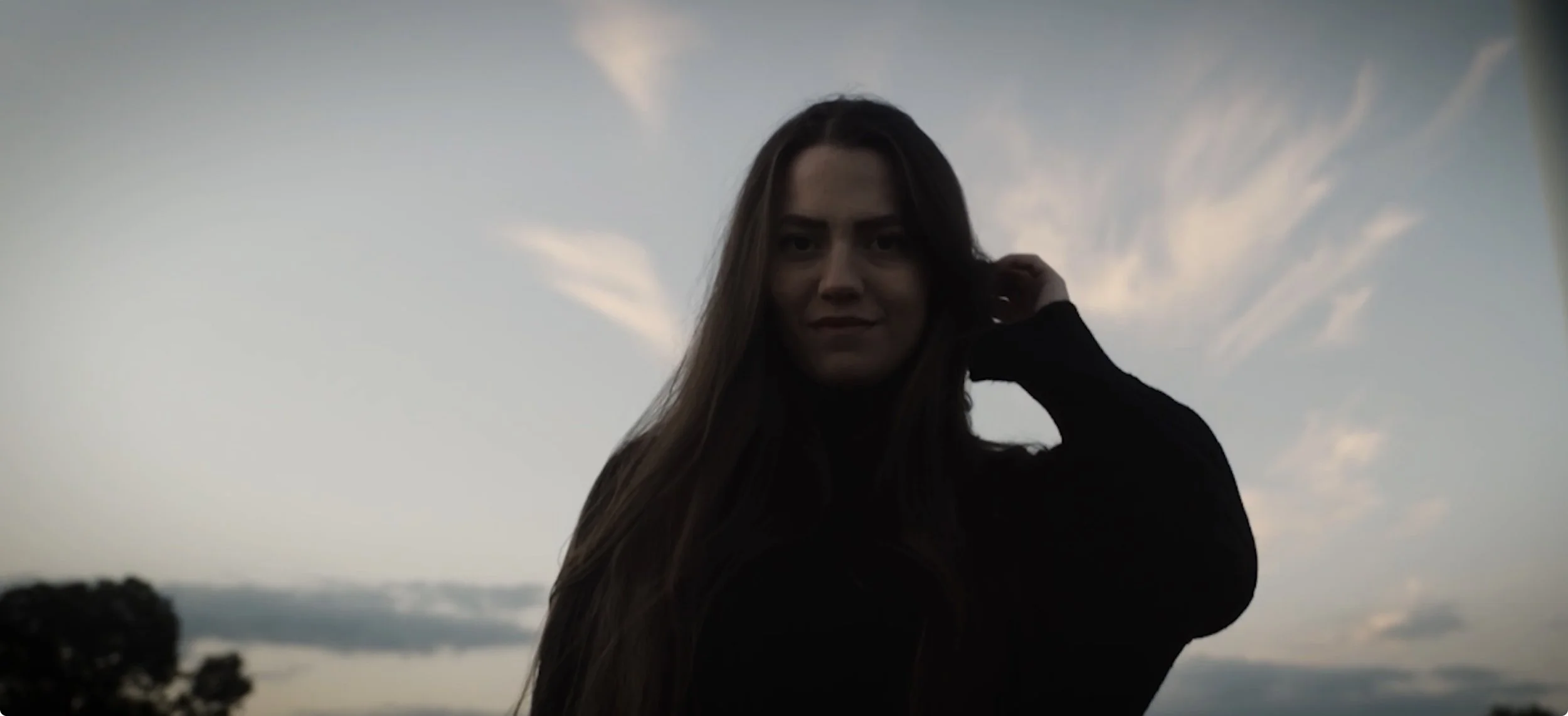 Cinematic Video A young woman with long brown hair, wearing black clothing, stands against a cloudy sunset sky and looks directly into the camera.