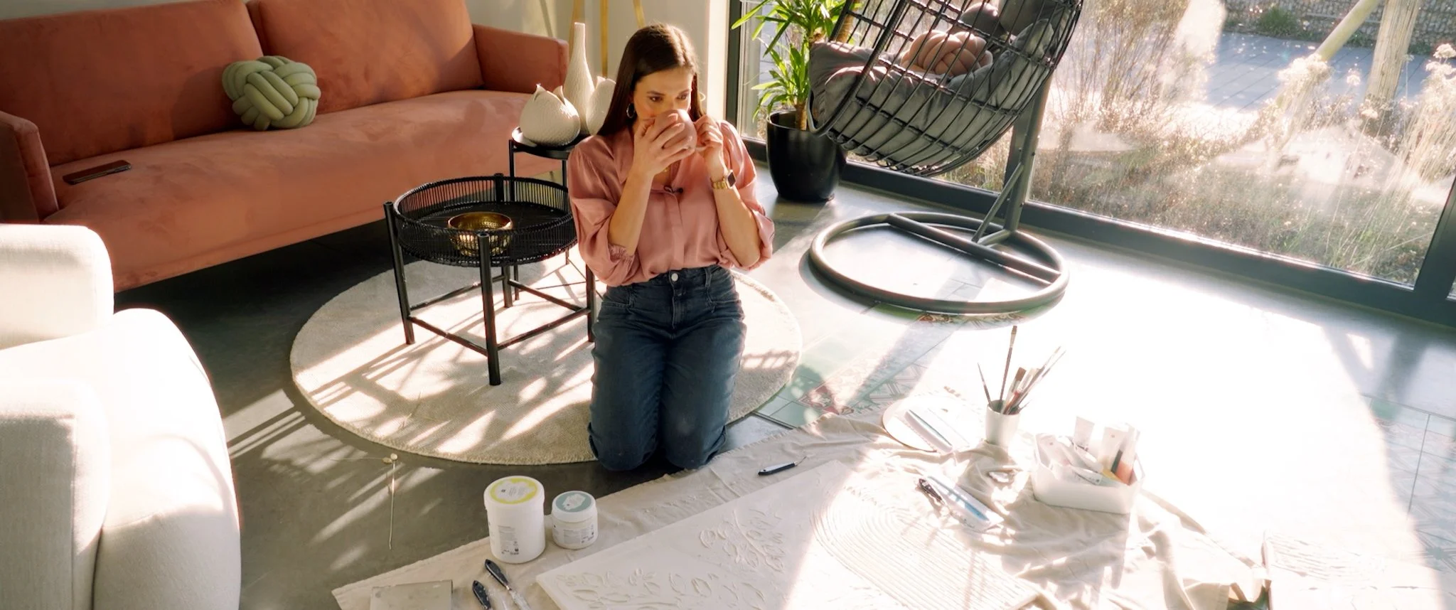 Eine Frau sitzt auf dem Boden in einem modern eingerichteten Wohnzimmer, hält eine Tasse in der Hand und scheint nachdenklich zu sein. Im Hintergrund sind große Fenster mit Blick ins Freie, ein Schlafsessel, eine Couch und einige Dekorationsgegenstände zu sehen. Im Vordergrund liegen Malutensilien und bei den Füßen der Frau befinden sich Dosen mit Farben.