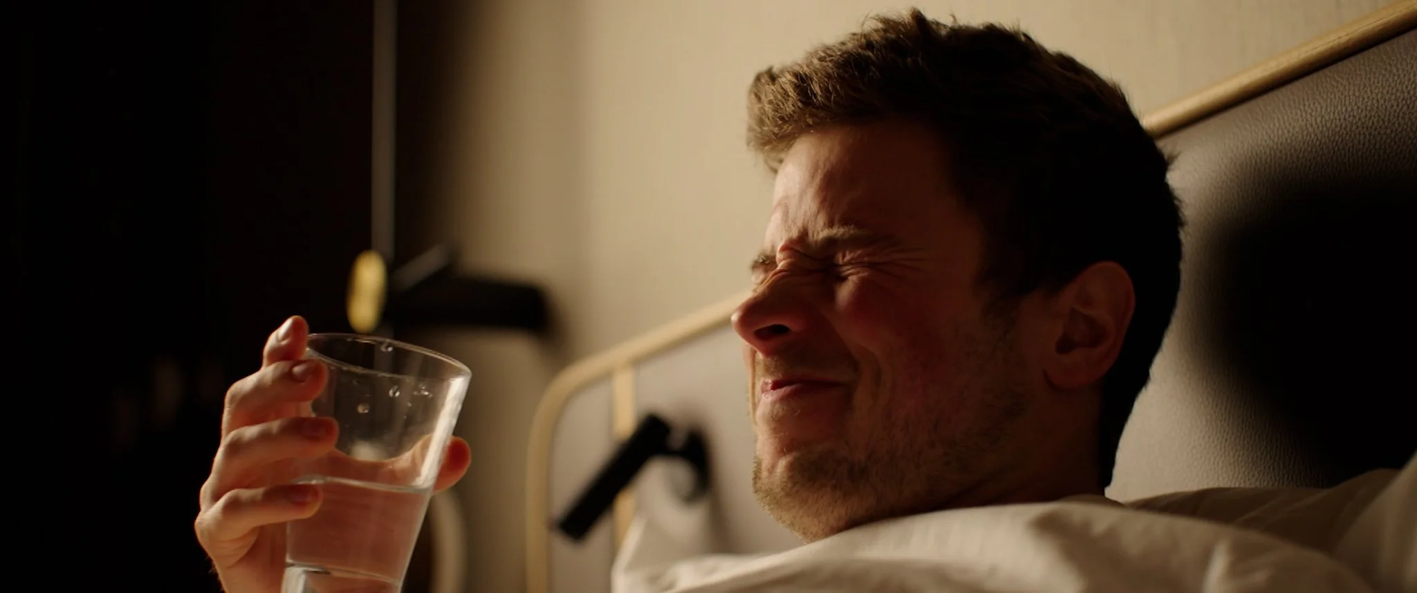 Cinematic Video, Short Film A man lies in a hospital bed, holding a glass of water and appearing to be in pain.