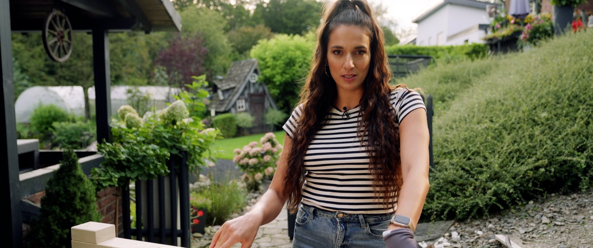 Cinematic Video, Sallys Welt, Saliha Özcan, A young woman with long, curly brown hair wears a black-and-white striped T-shirt and a smartwatch. She stands in a garden in front of a fence surrounded by plants and flowers, with houses and trees visible