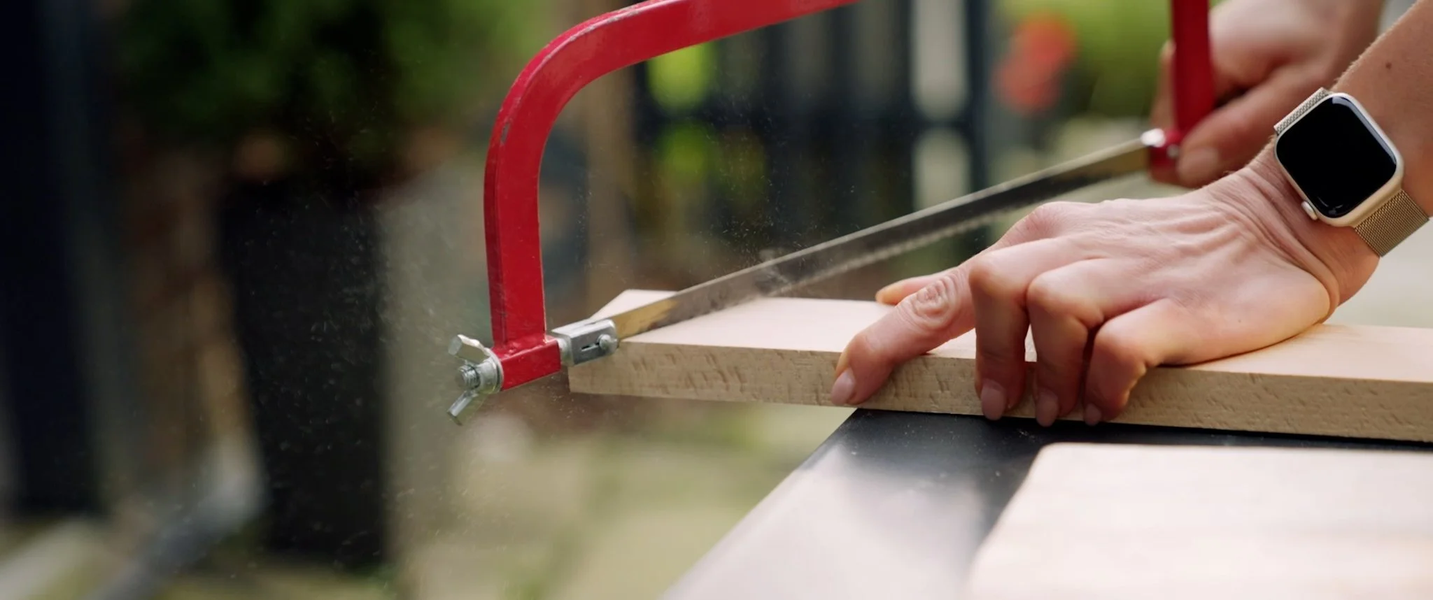 Cinematic Video Close-up of a person cutting a piece of wood with a hand saw. The person is wearing a smartwatch, with a garden visible in the background.