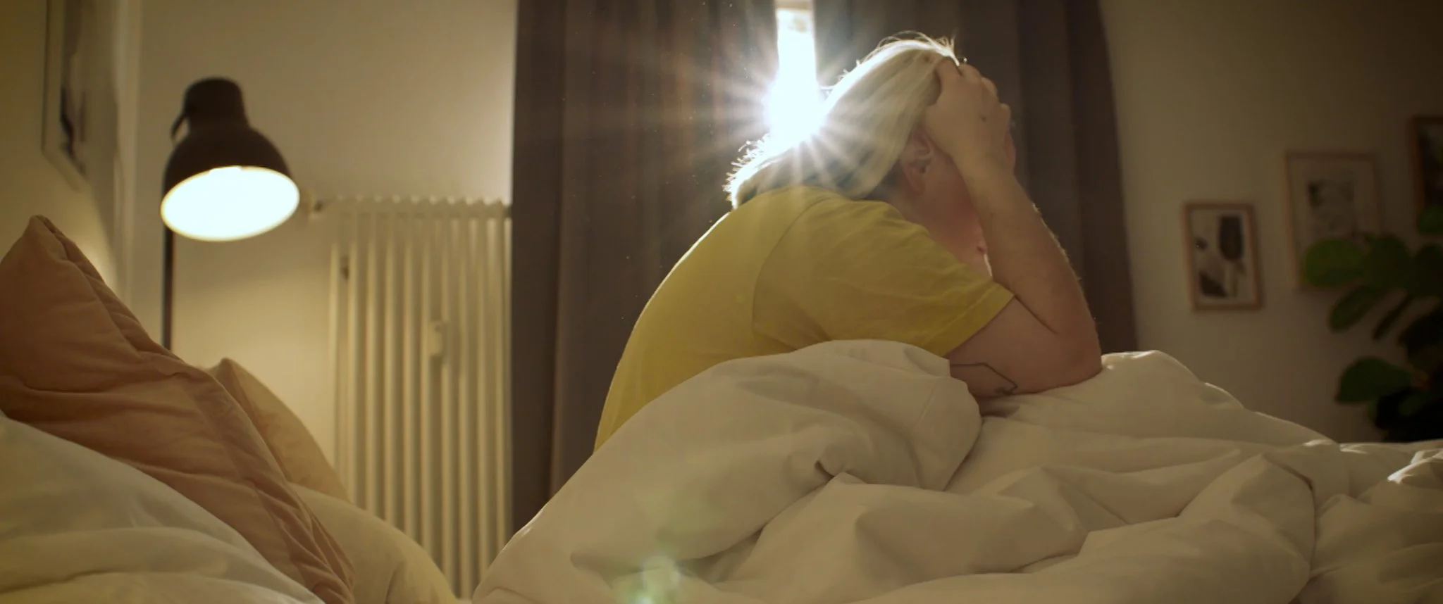 Cinematic Video, SHort Film, An older person sits distressed on a bed in a sunlit bedroom, holding their head in their hand.
