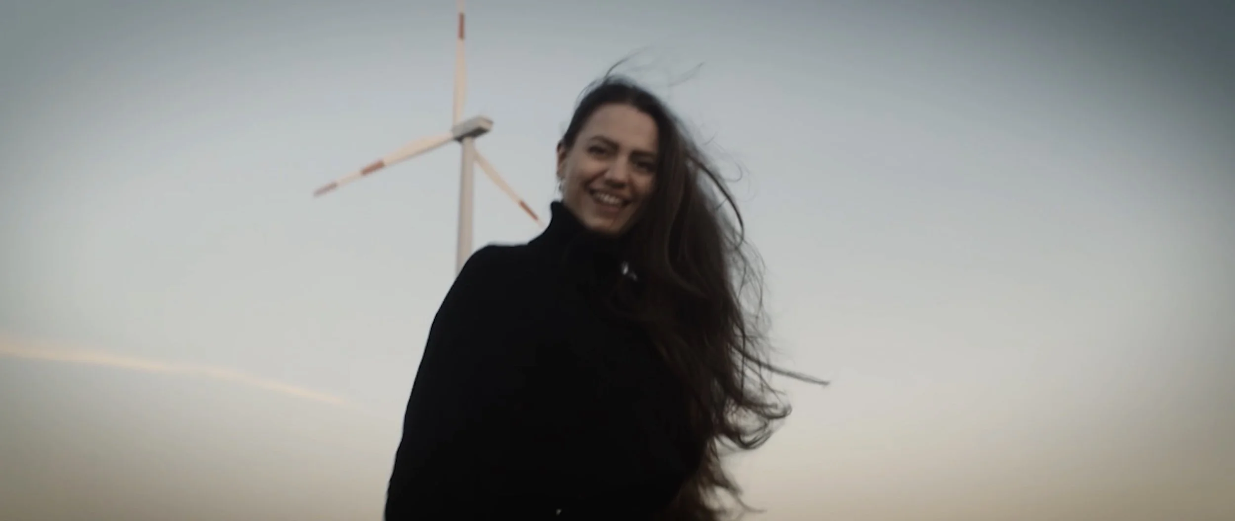 Cinematic, Showrt Film, A laughing woman with long, windblown hair stands in front of a wind turbine at dusk.