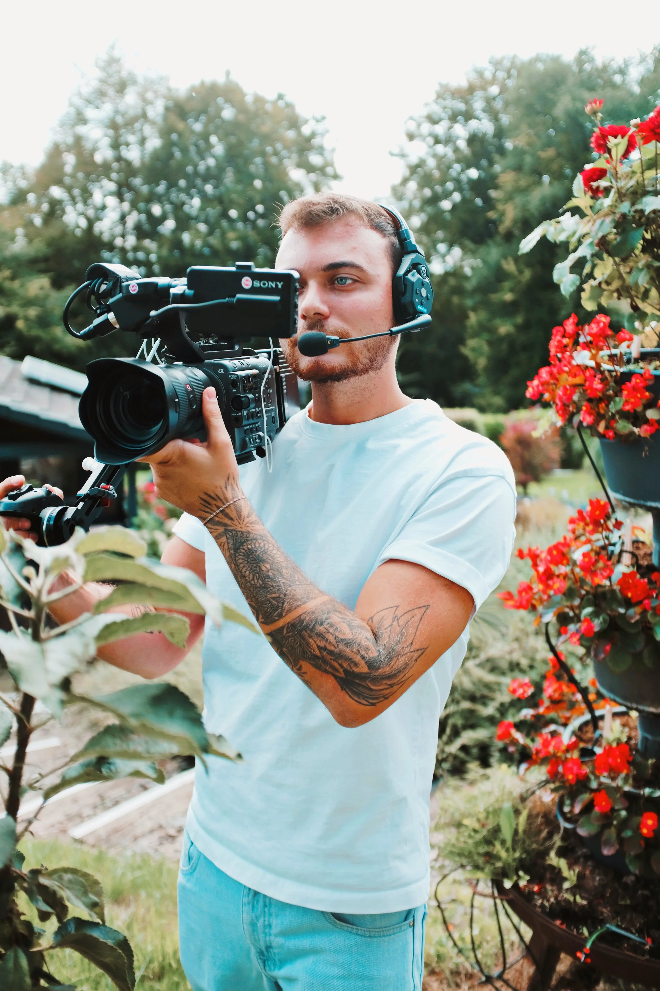 Junge Mann mit Kamera und Headset fotografiert Pflanzen im Garten, umgeben von Blumen und Bäumen.