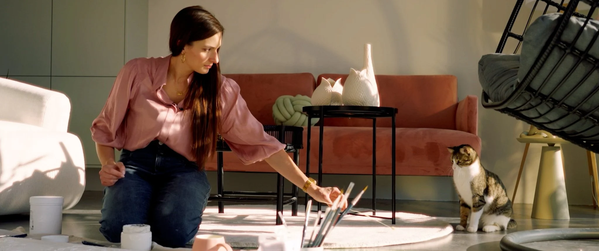 Frau in Wohnzimmer mit Katze, bei kreativer Aktivität, im Hintergrund Sofa, Kissen und dekorative Gegenstände auf Tisch, natürlicher Lichtschein, entspannte Atmosphäre.