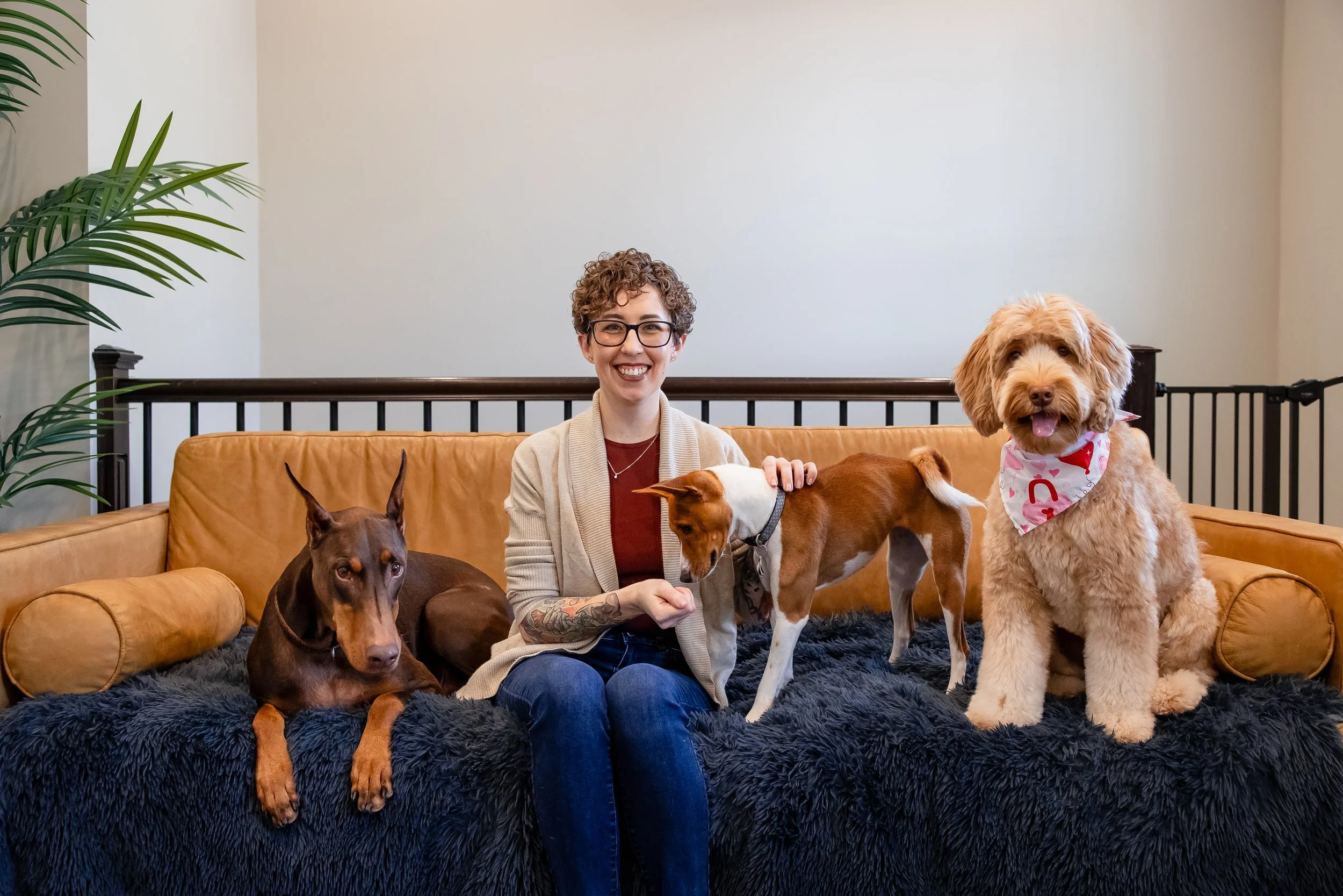 Smiling woman sitting on a couch with three dogs, including a large goldendoodle wearing a pink bandana.