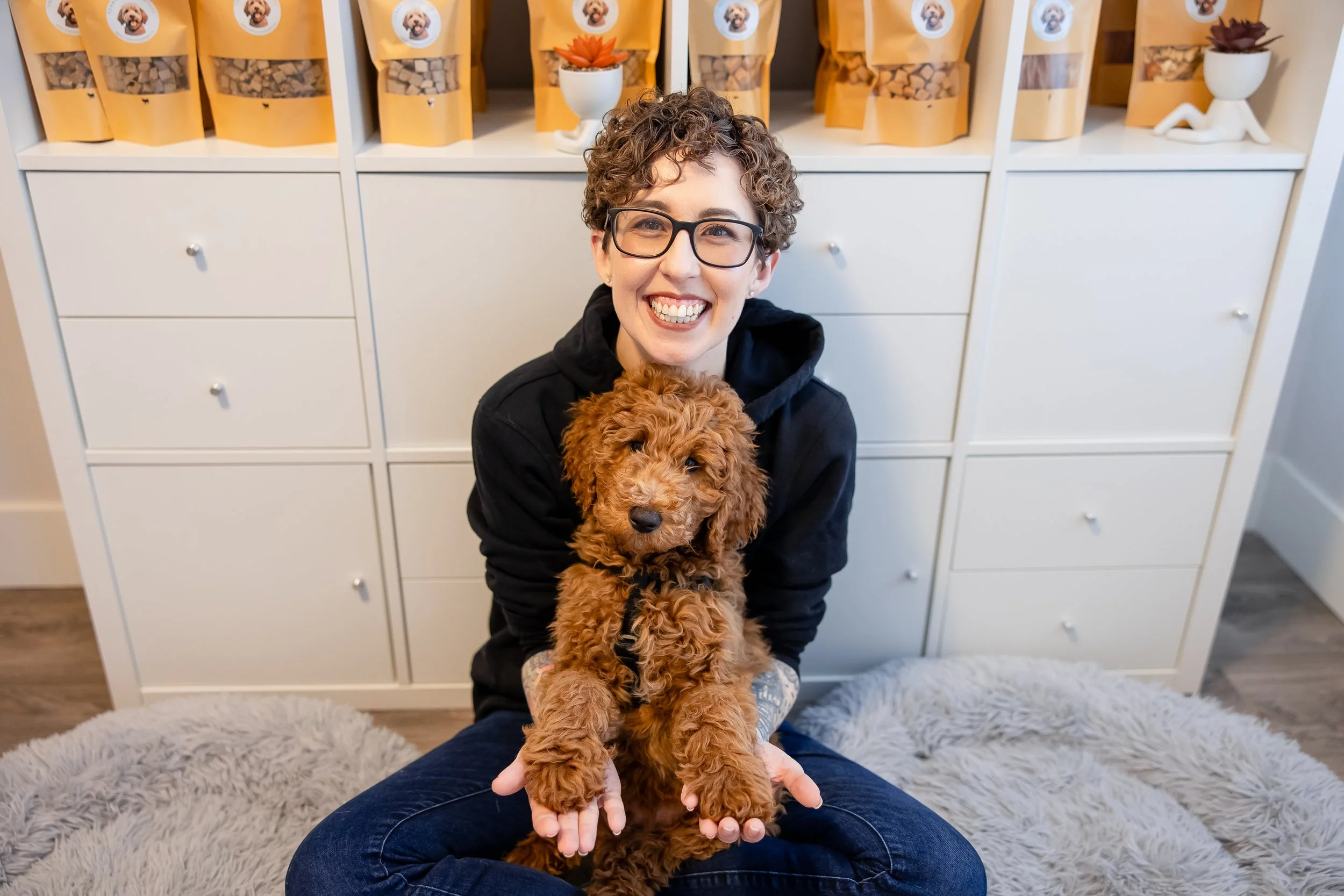 Smiling person holding a curly-haired brown puppy in front of dog treat bags and home decor