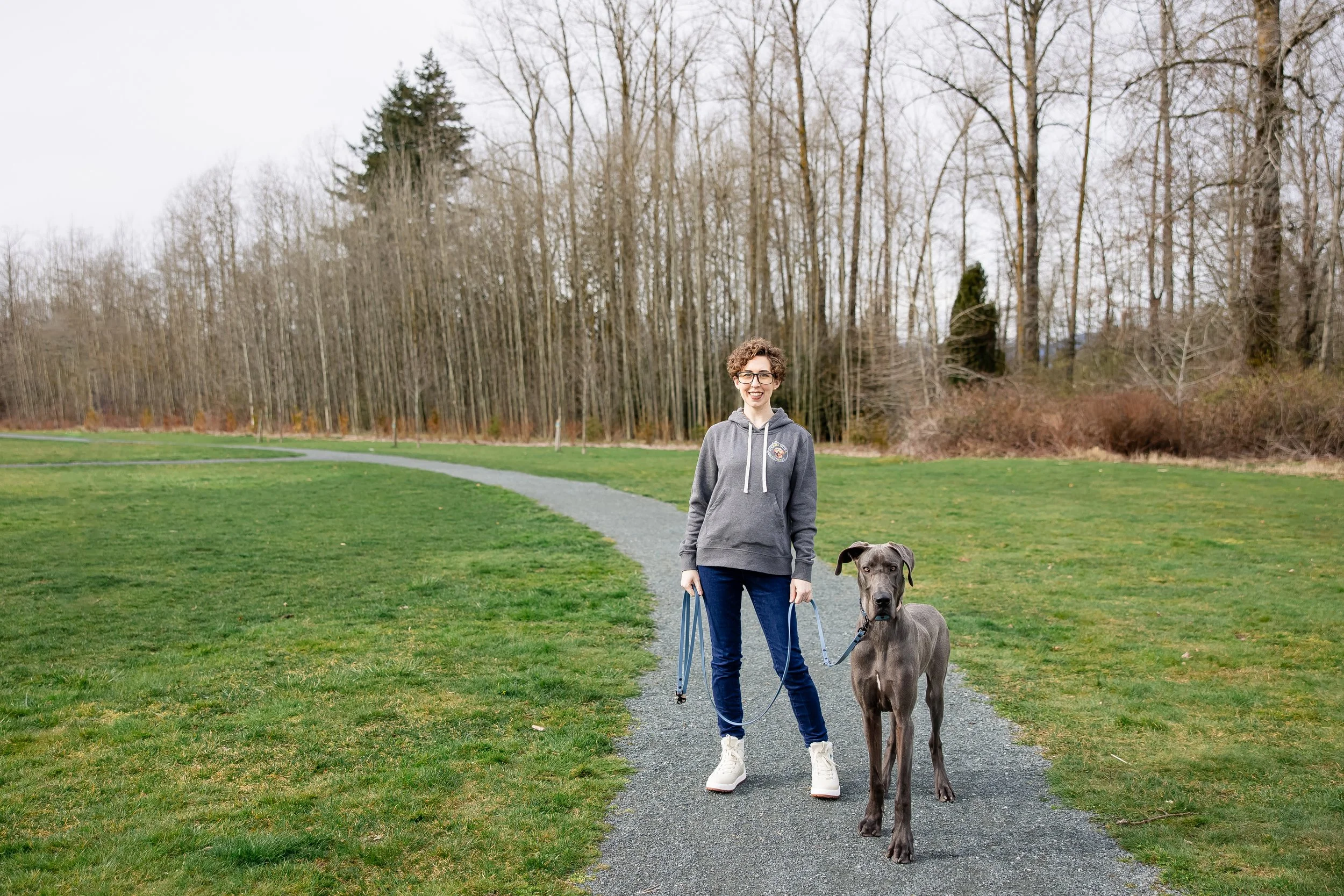 Person walking a Great Dane on a leash along a paved park trail surrounded by grass and leafless trees