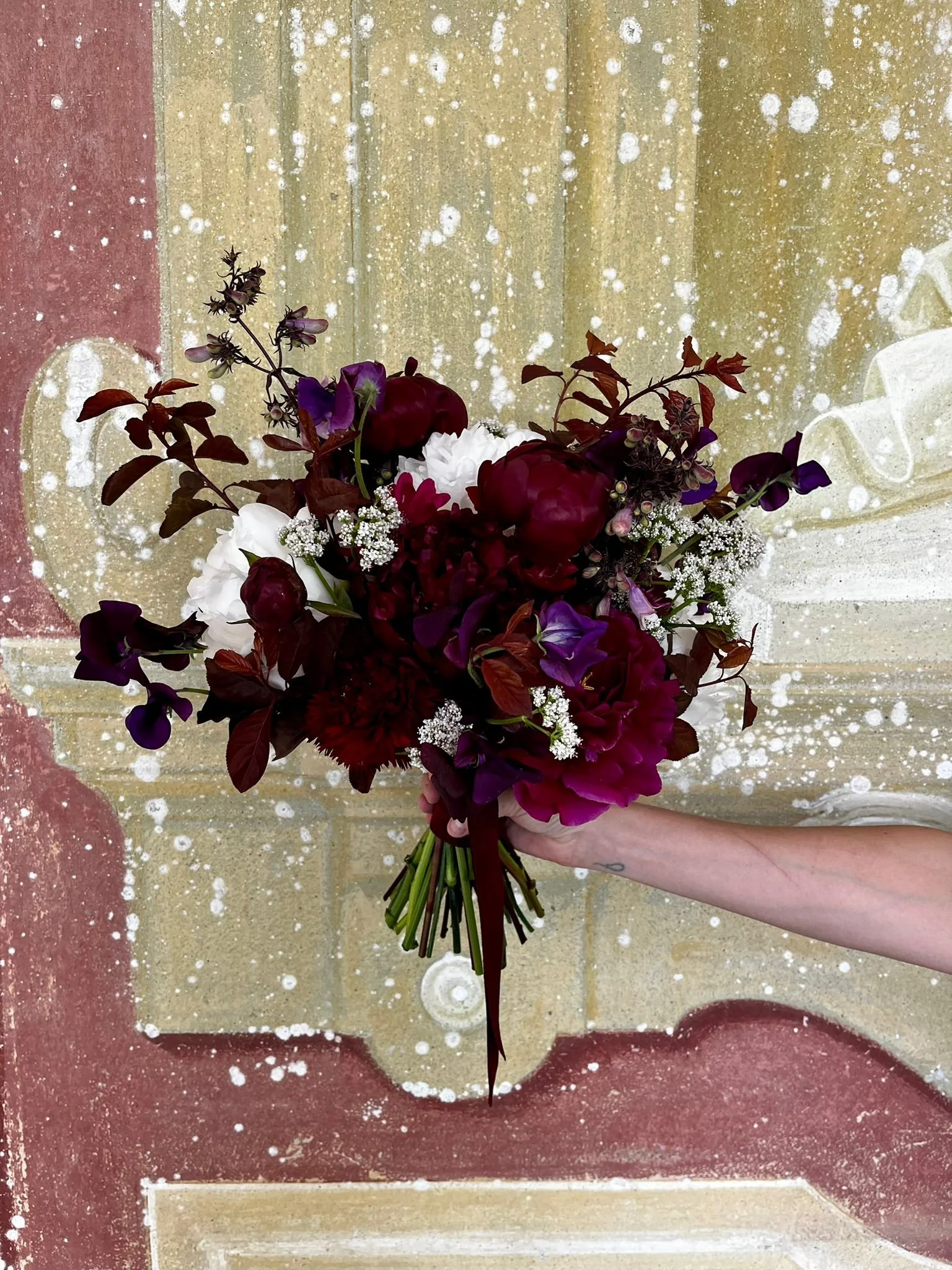 A person holding a bouquet of mixed flowers against a painted concrete wall with white specks.