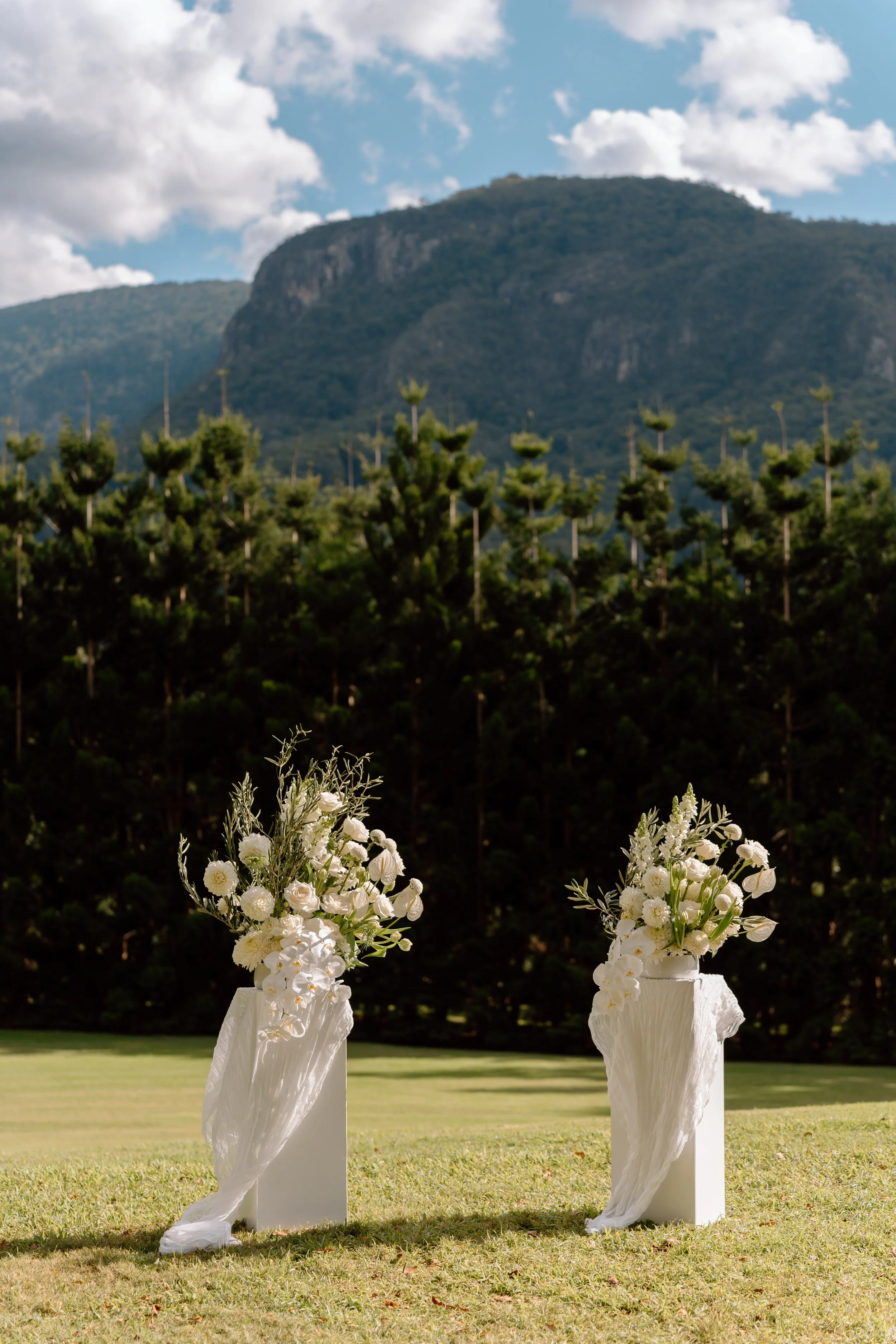 Two wedding flowers arrangements on white pedestals with a grassy outdoor setting and trees and a mountain in the background.