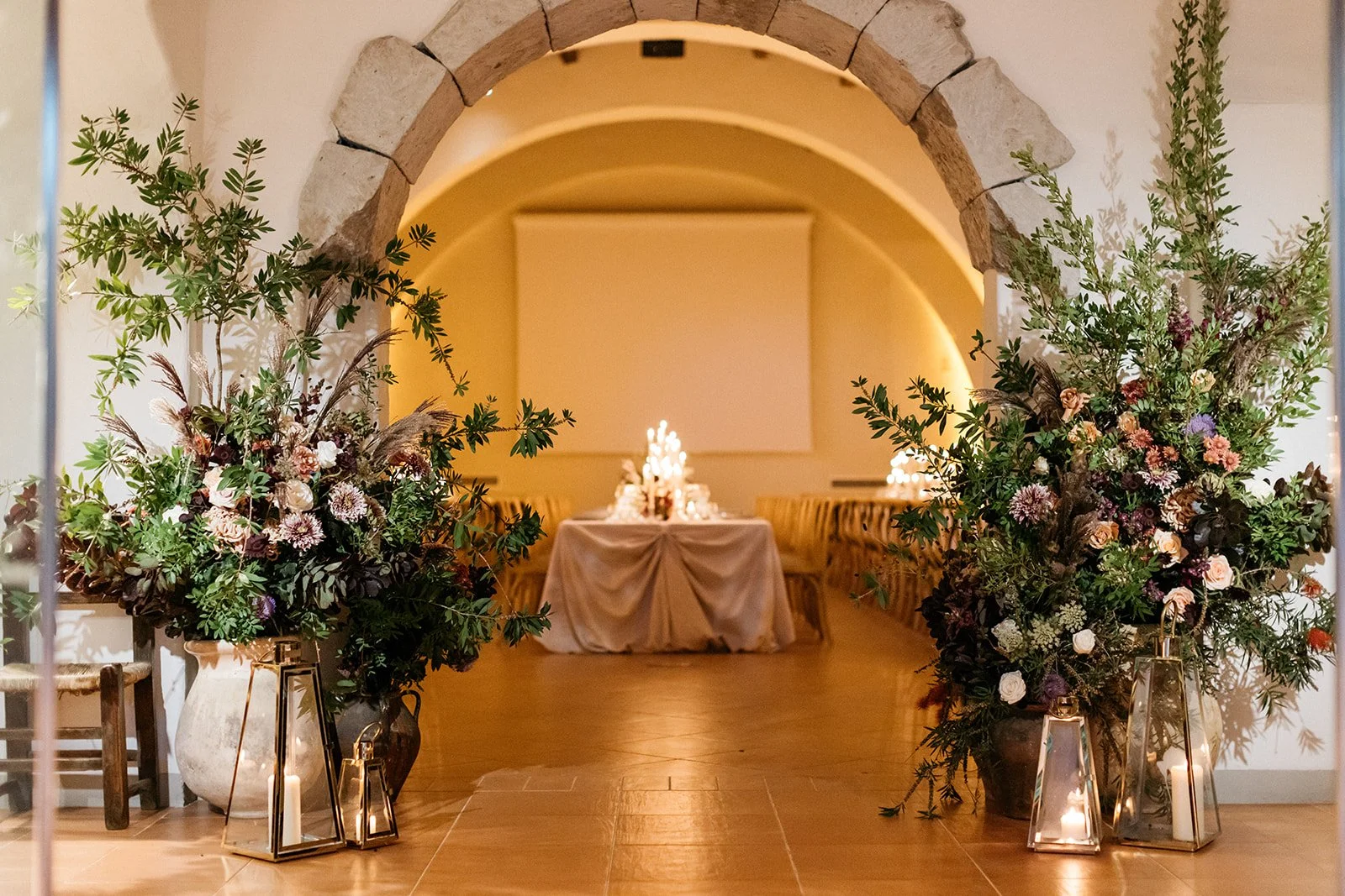 A decorated reception area with large floral arrangements on either side of an arched doorway, leading to a table with candles and floral centerpieces inside a warmly lit room. Su Gologone Sardinia, Sardegna