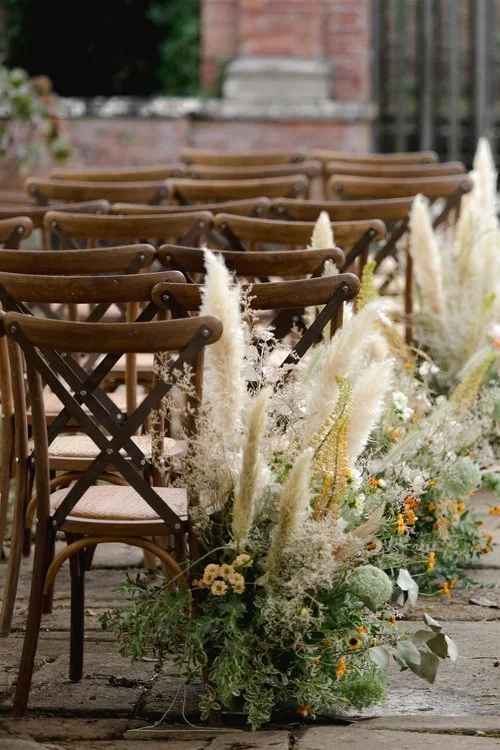 Wooden chairs lined up outdoors next to floral arrangements with pampas grass and white flowers. Villa Buonvisi Lucca